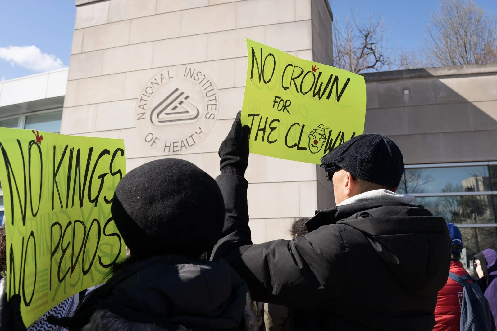 Couple holding protest signs saying "No Kings, No Pedos" and "No Crown for the Clown"