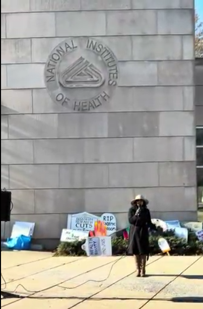 A person speaking at an NIH vigil, with the NIH logo visible on the wall above thema