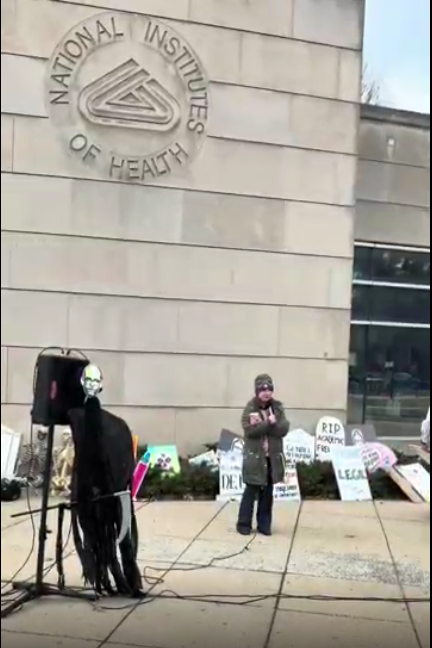 A person speaks into a microphone at an NIH vigil
