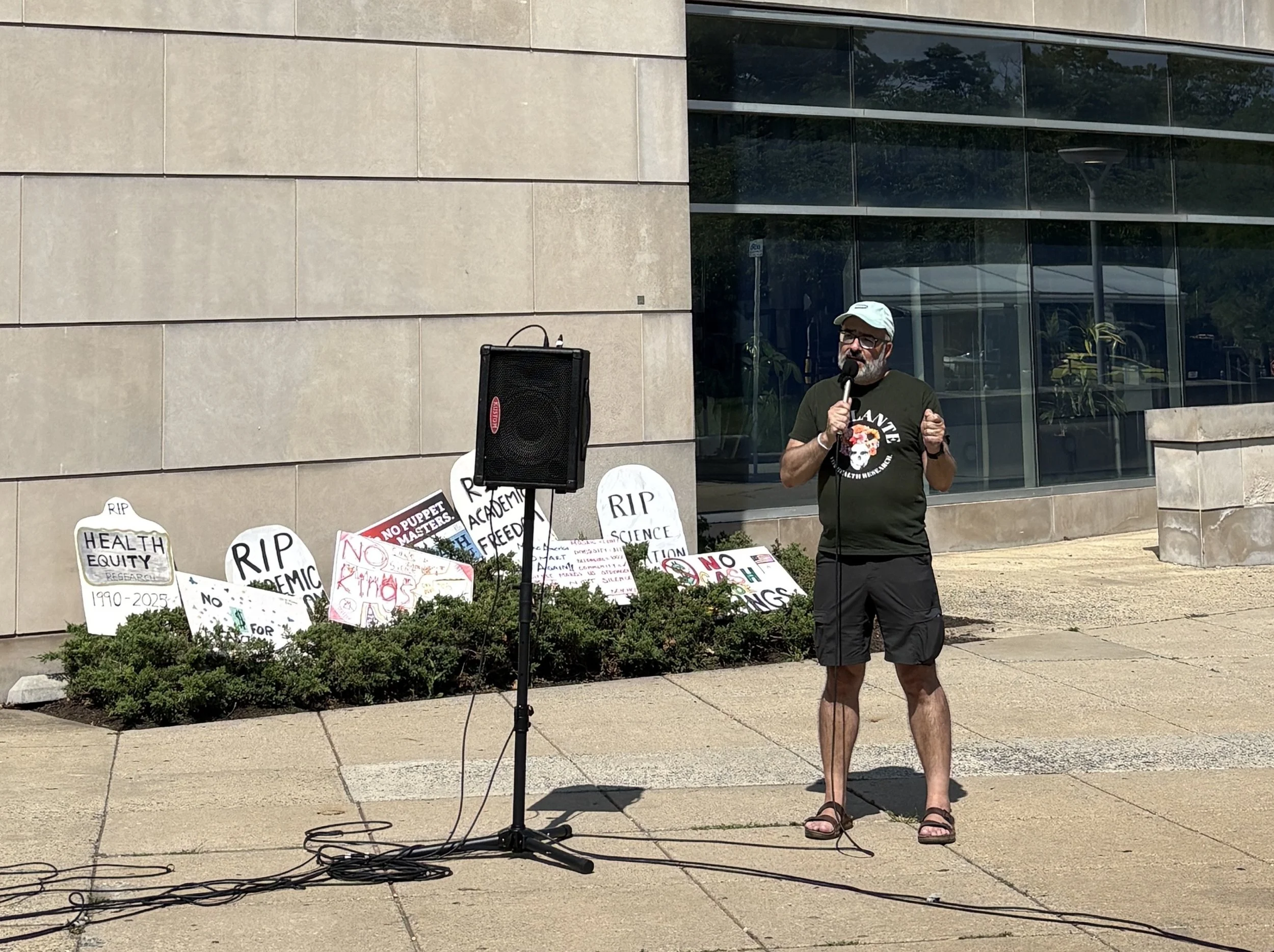 A man speaks into a microphone at the NIH Vigil with protest signs displayed behind him.
