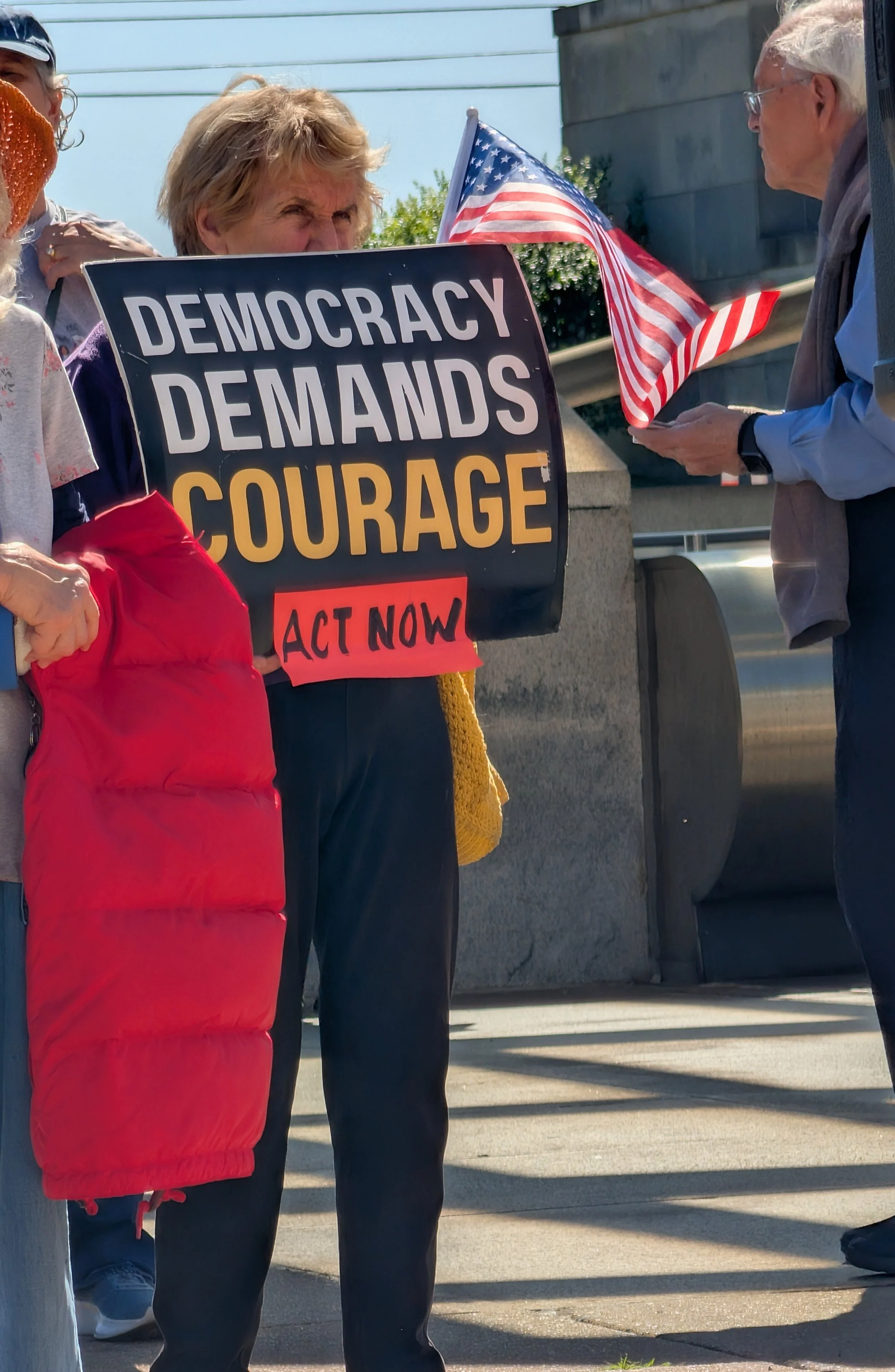 A woman watching the vigil holds an American flag and a "Democracy Demands Courage - Act Now" protest sign