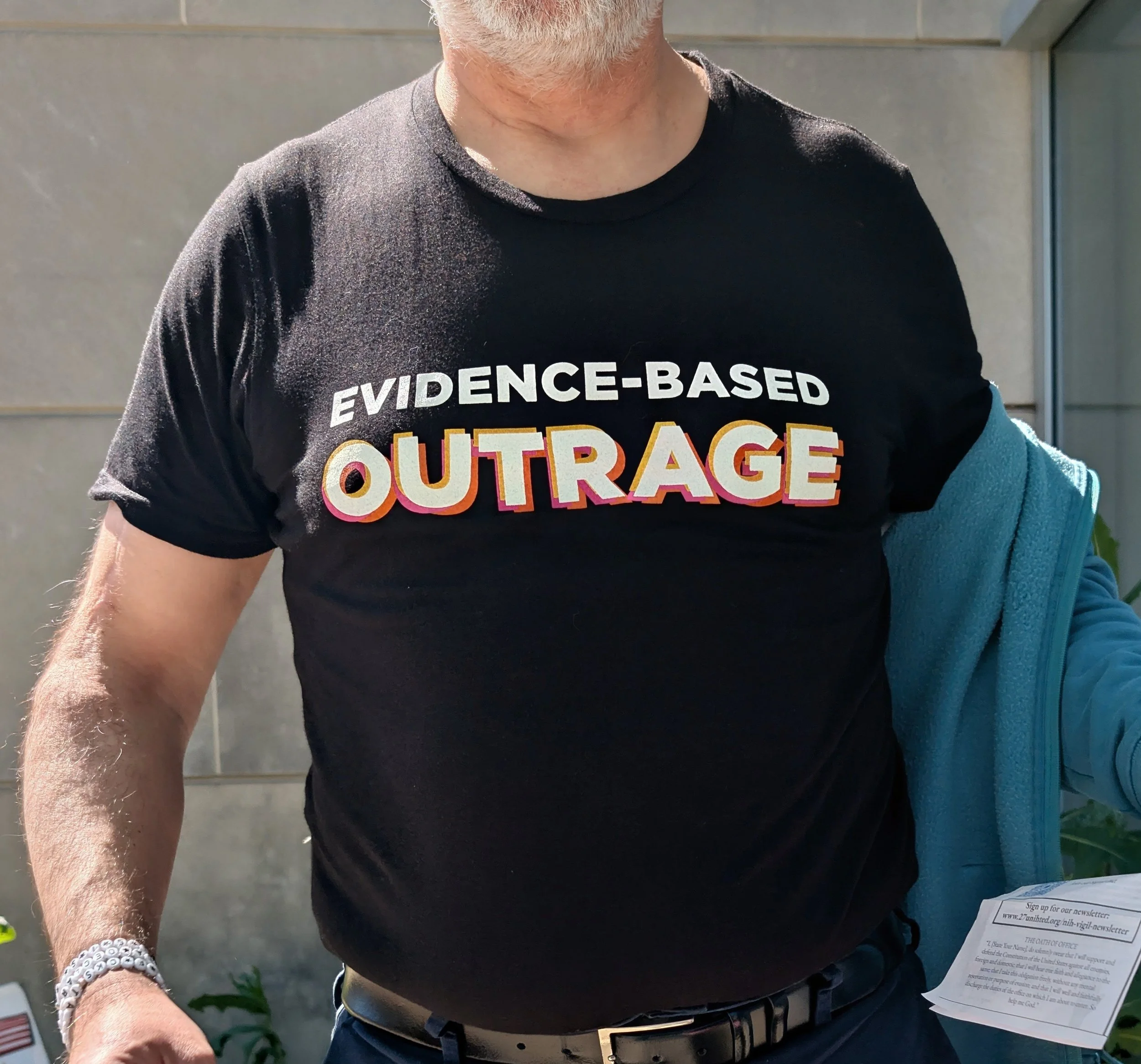 A man holding a vigil program and wearing 27 UNIHTED friendship bracelets and a T-shirt that says "Evidence-Based Outrage"