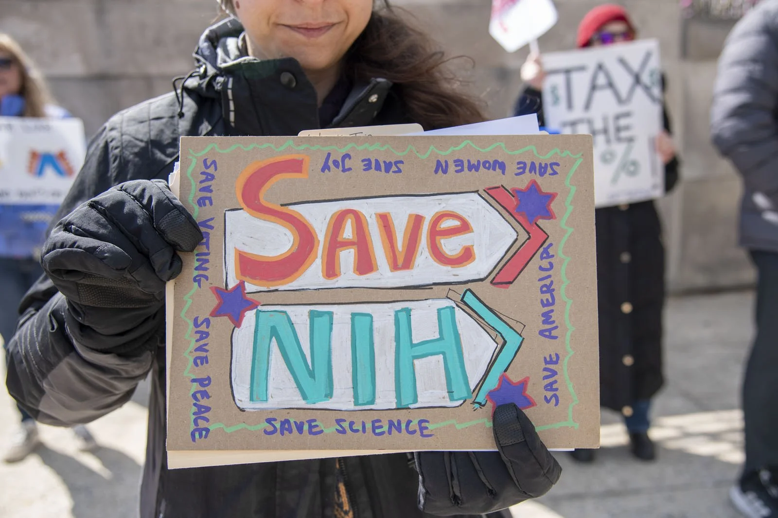A person holding a protest sign that says "Save NIH, Save America, Save Science, Save Peace, Save Joy, Save Women, Save Voting"