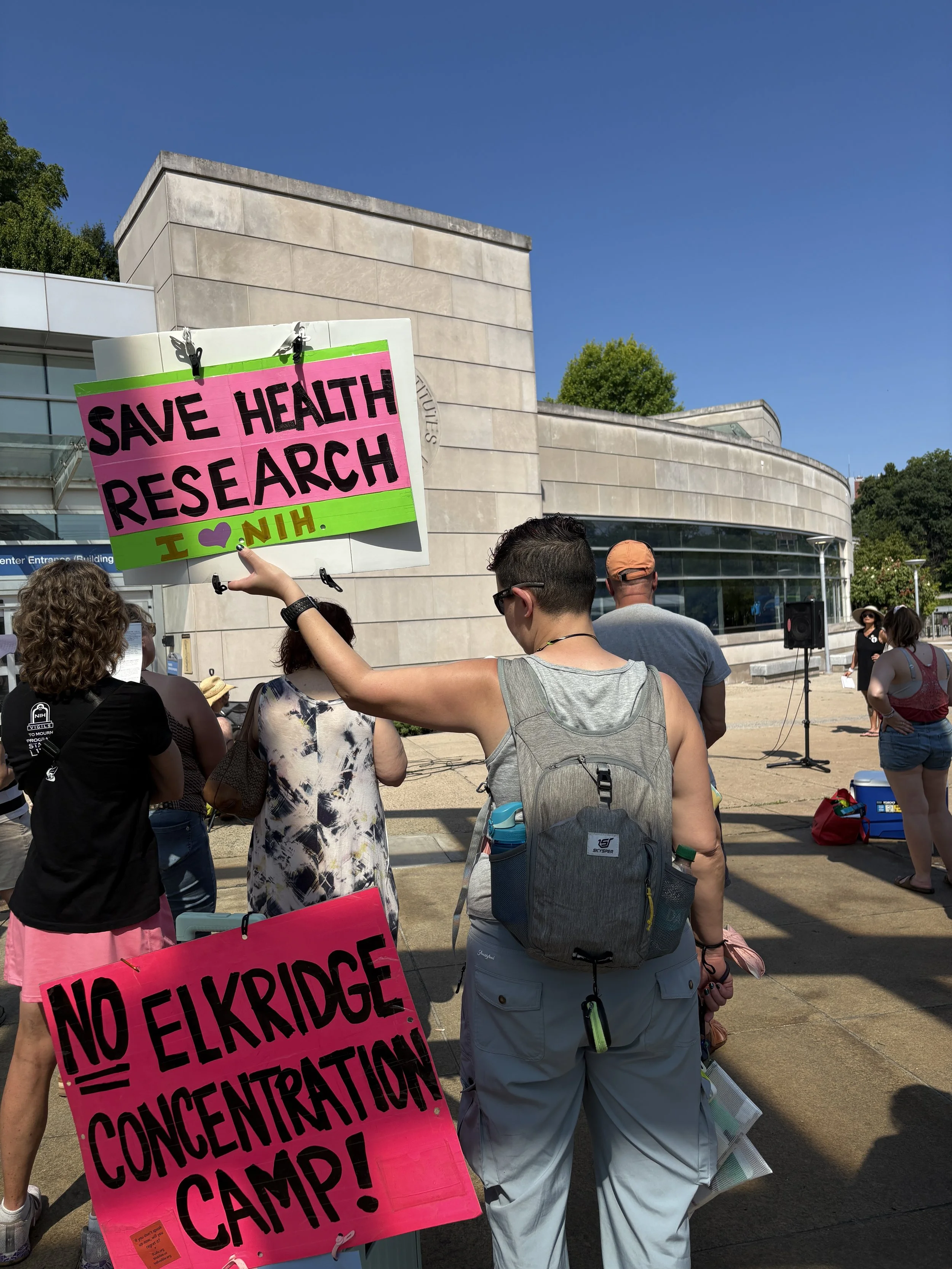 An NIH Vigil attendee holds a “Save Health Research” sign near another sign opposing Elk Ridge concentration camp.