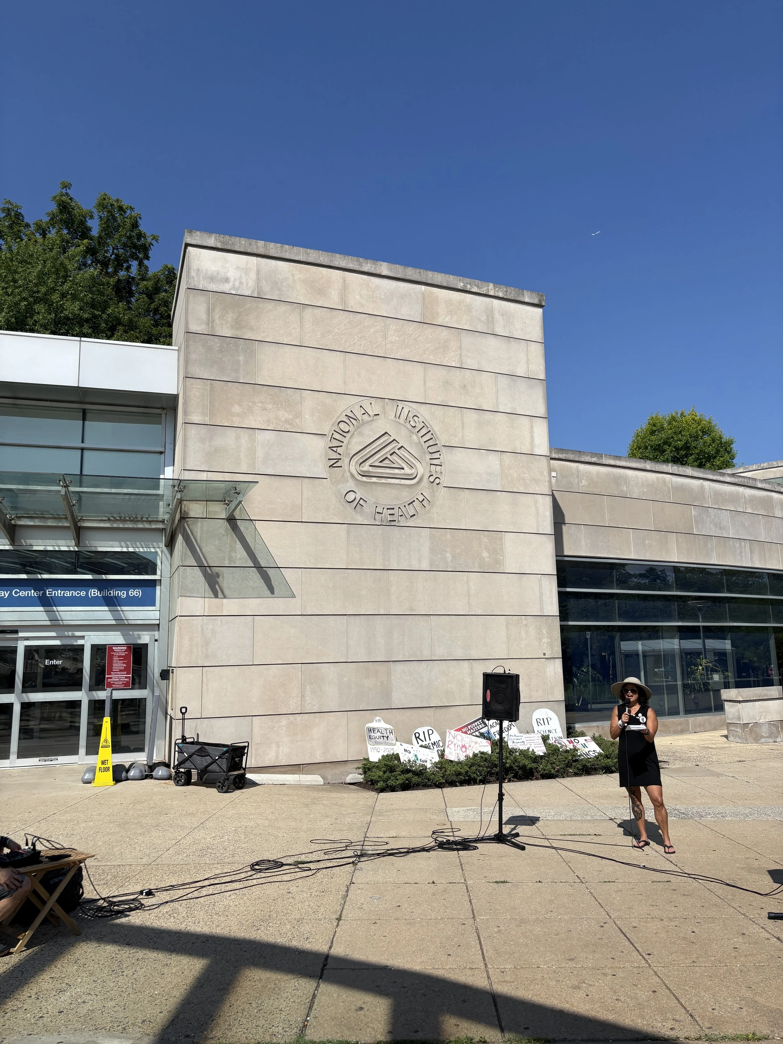 An NIH Vigil speaker stands outside the NIH Gateway Center near protest signs and audio equipment.