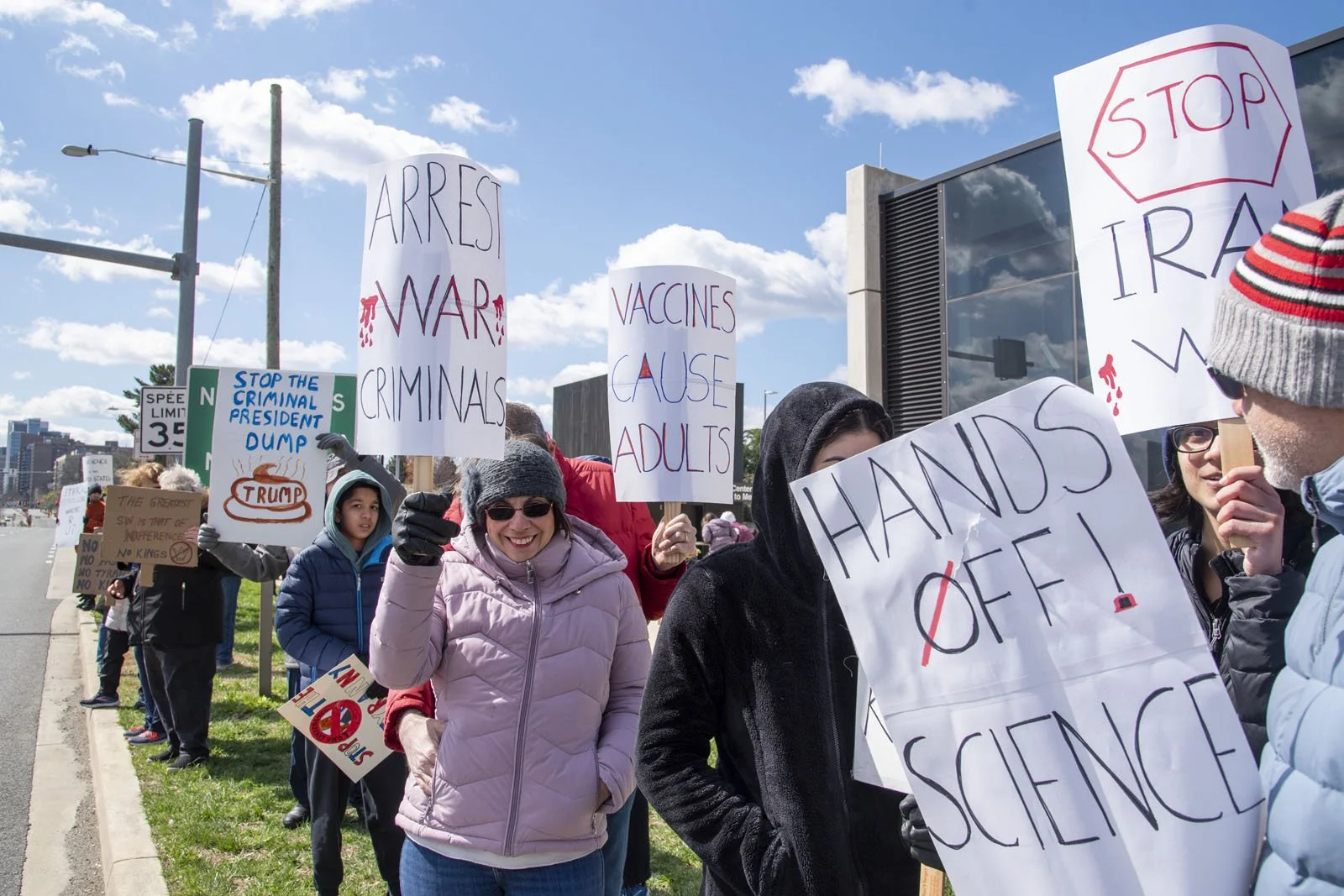 A group of roadside protestors holding signs saying "Arrest war criminals," "vaccines cause adults," and "hands off science"