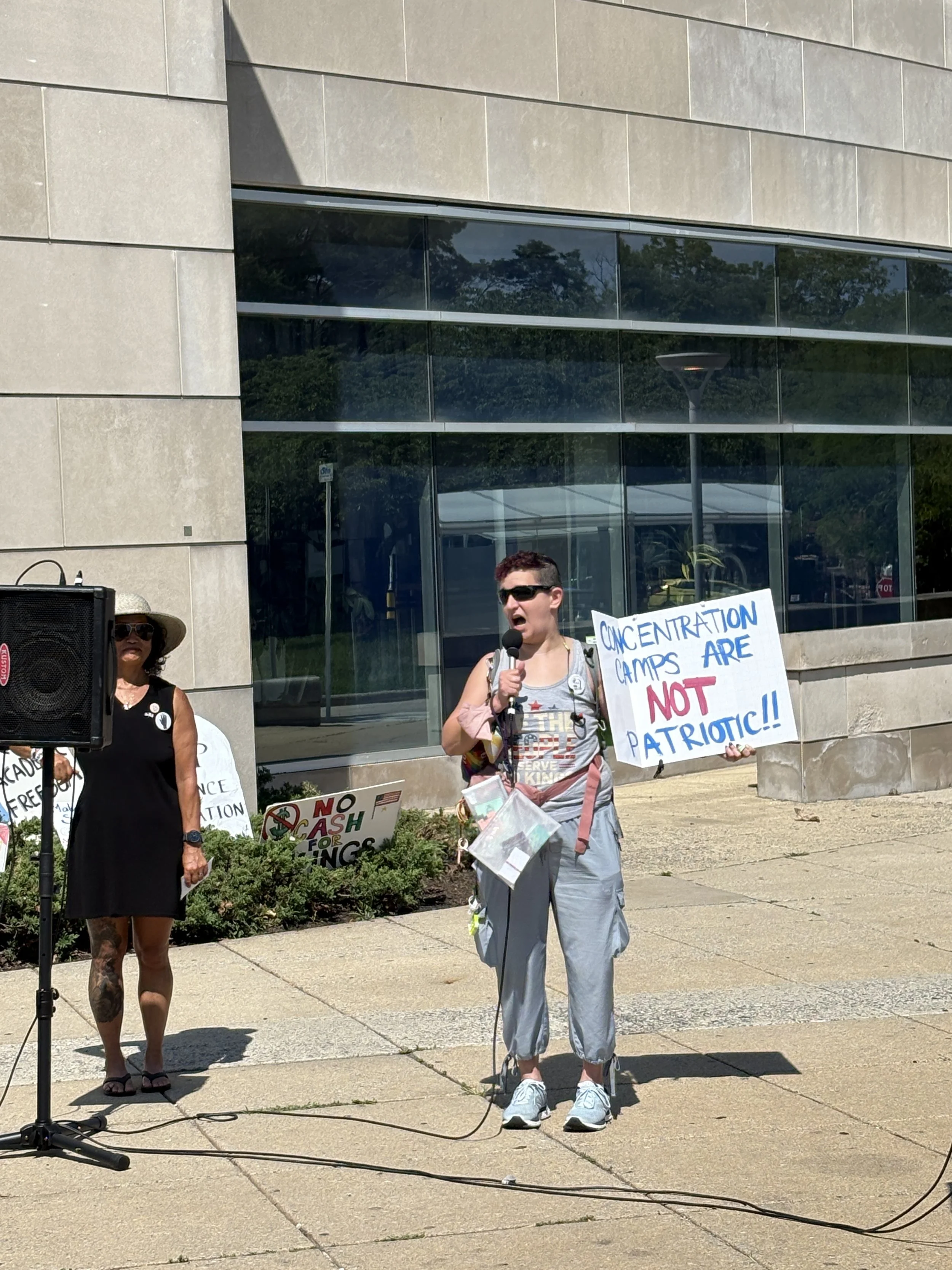 An NIH Vigil attendee speaks into a microphone while holding a sign that reads “Concentration camps are not patriotic!!”