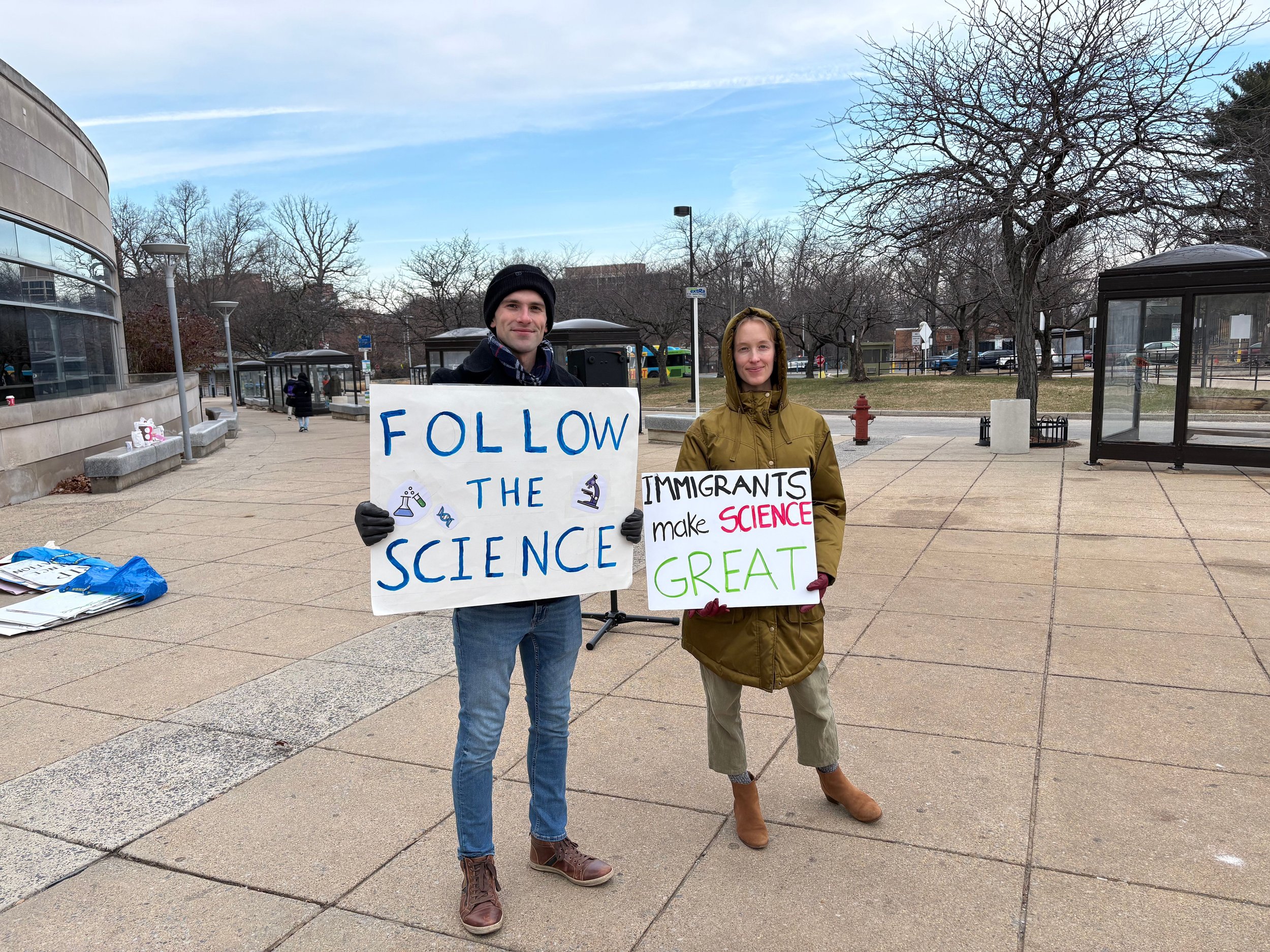 A couple holds protest signs reading "Follow the science" and "Immigrants make science great"