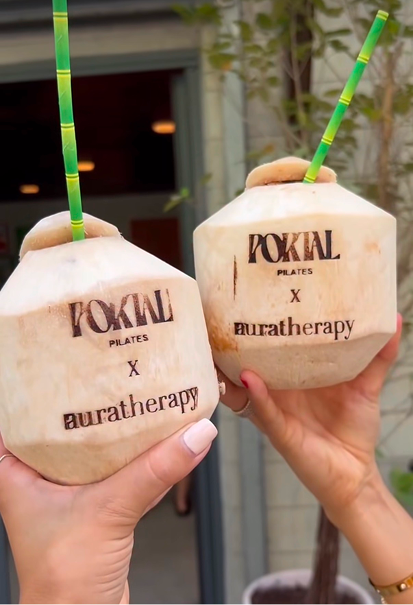 Two coconuts with straws and lids, each branded with 'Portal Pilates', being held up by two hands in front of a building.
