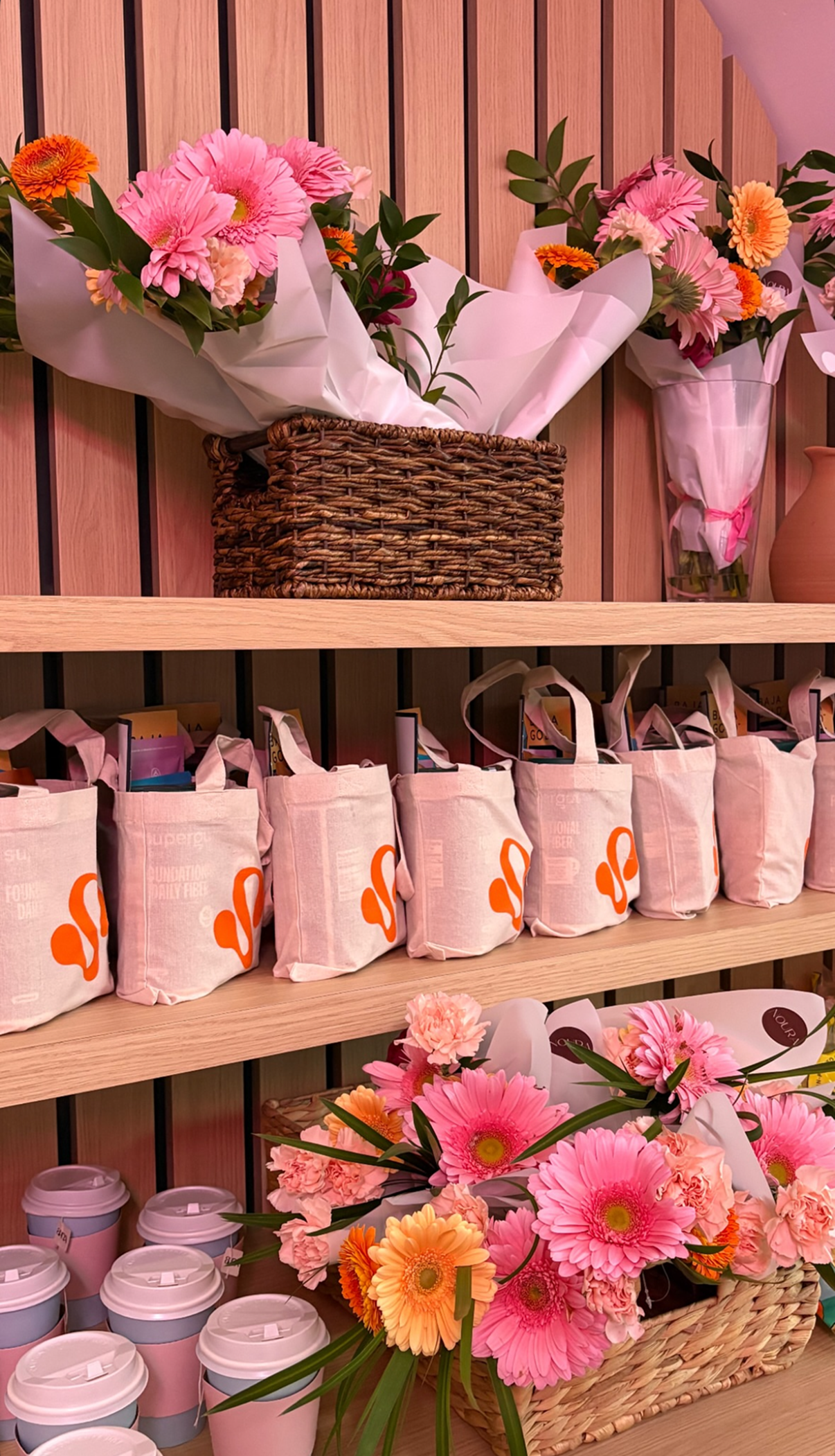 Display of pink, orange, and peach flower bouquets, pink tote bags with orange logo, and pink and white coffee cups arranged on wooden shelves.