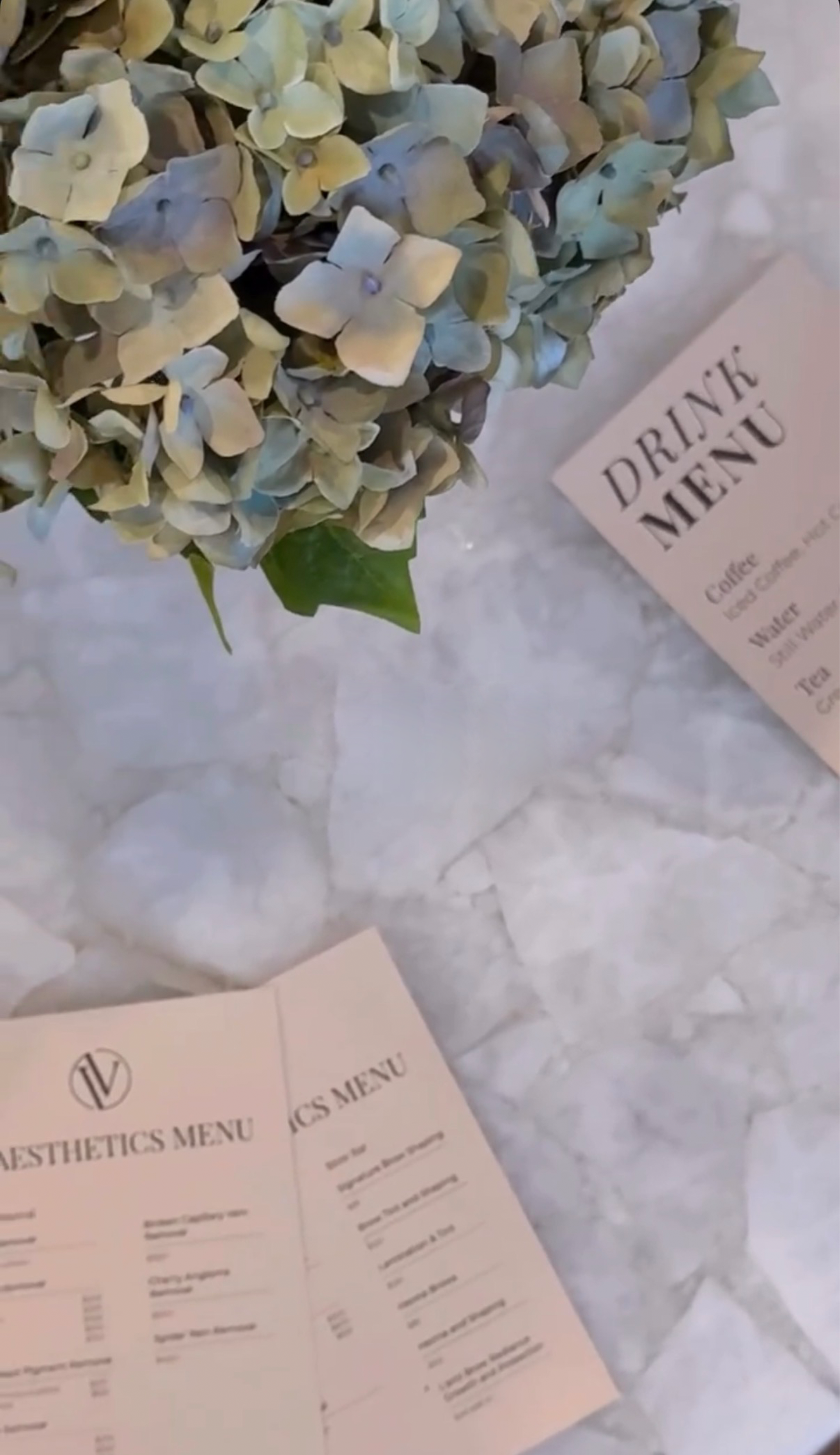 A bouquet of light-colored hydrangea flowers on a marble surface with a drink menu and an aesthetics menu nearby.