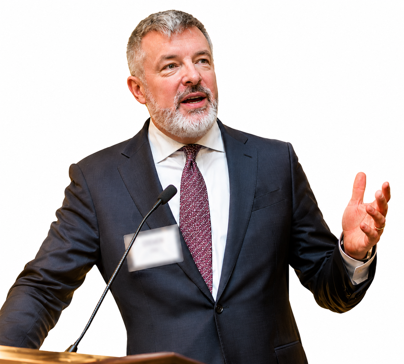 A man in a dark suit and tie is speaking at a podium with a microphone, gesturing with his right hand, with a white background.