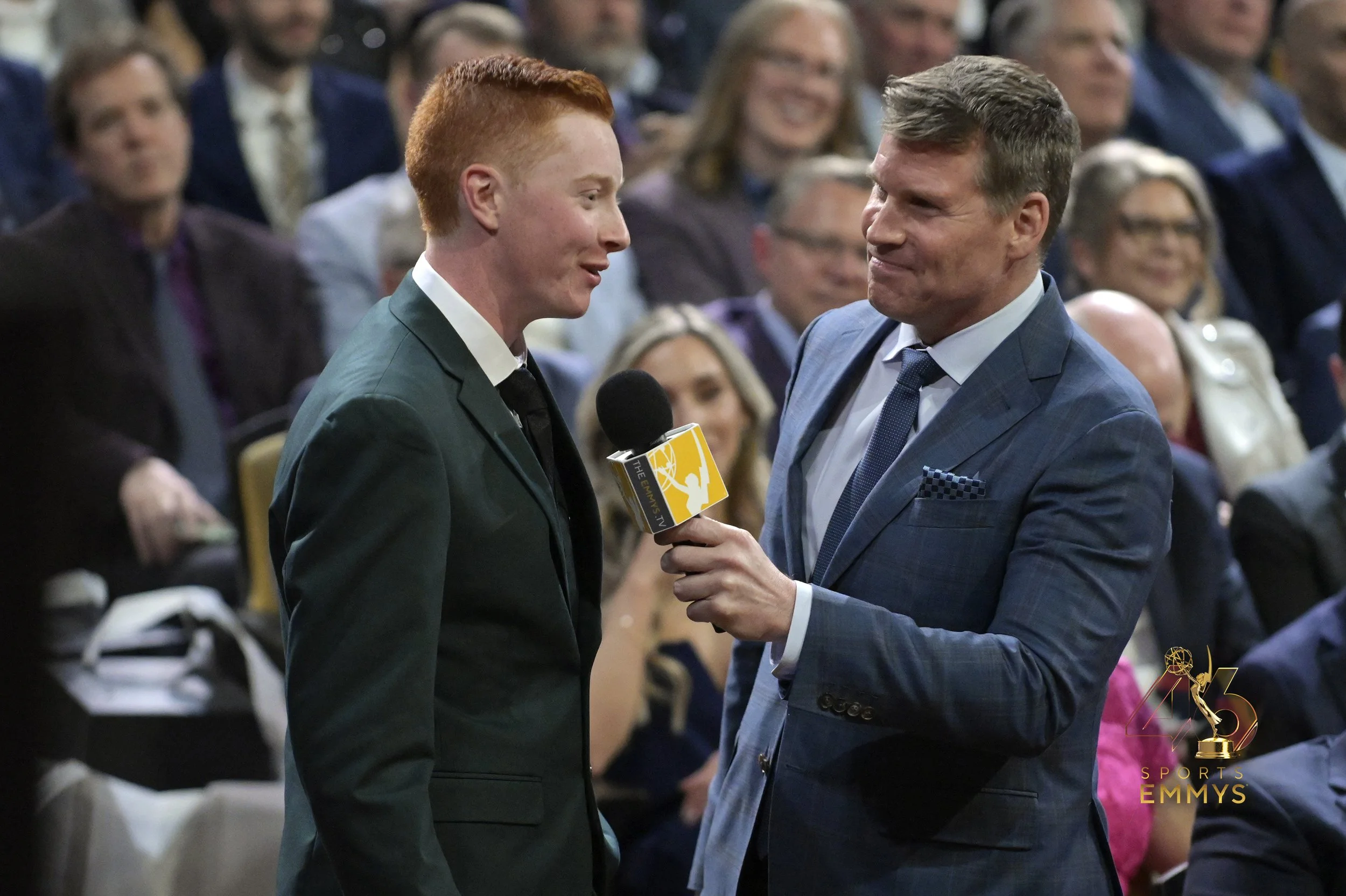 An interview at the sports Emmys showing a man in a blue suit holding a microphone and smiling, talking to a young man in a green suit. The audience in the background is seated and watching the interaction. Josiah Wonnell & Scott Hanson.