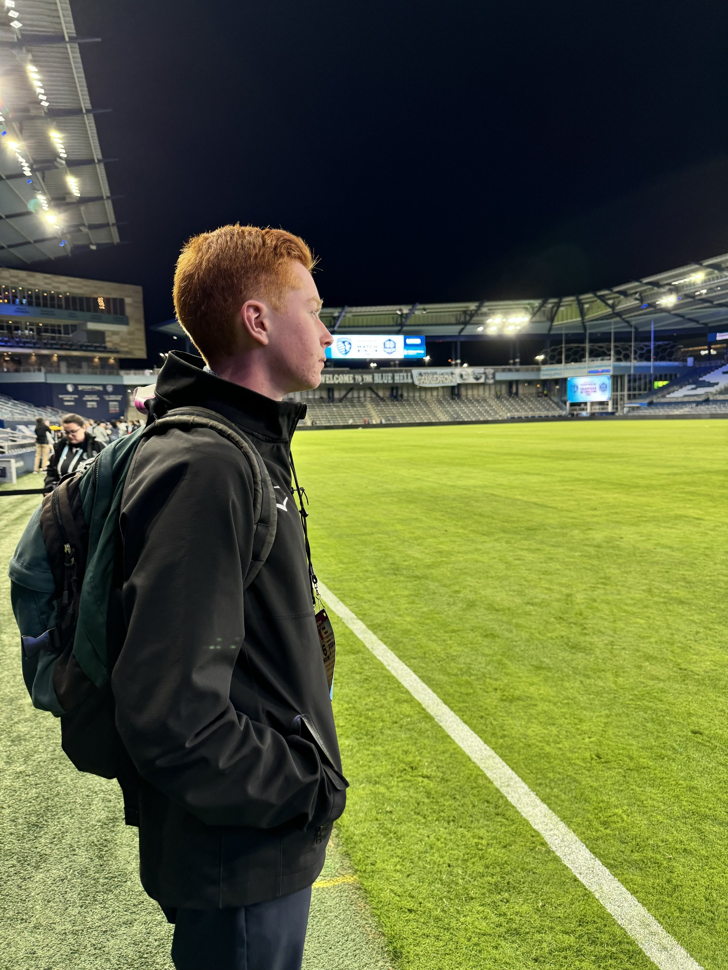 A young man with red hair wearing a black jacket and backpack standing near the edge of a soccer field at night, looking across the field at a stadium.