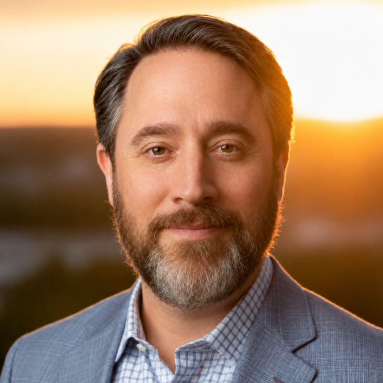 Close-up of a middle-aged man with brown hair and beard, wearing a checked shirt and blazer, outdoors during sunset.