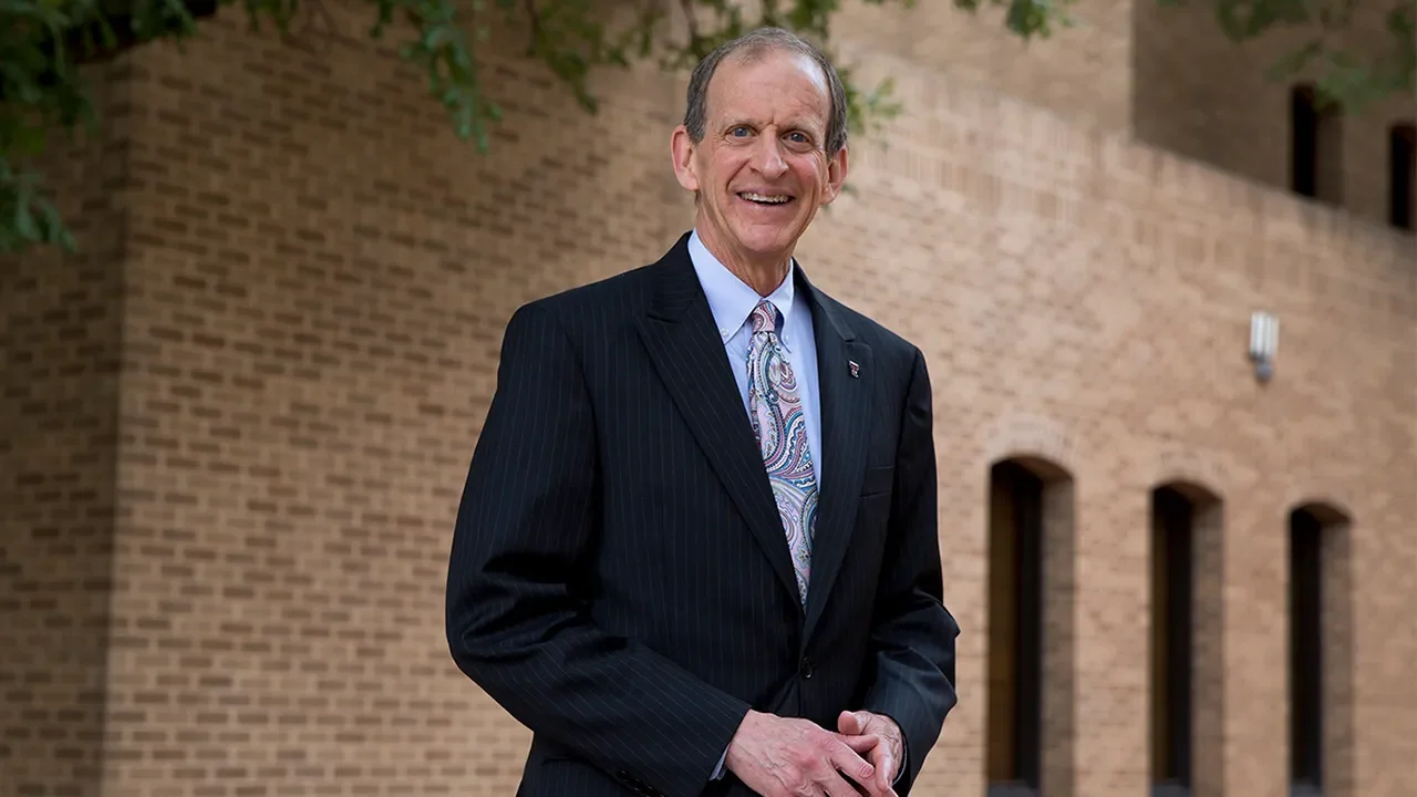 A smiling elderly man in a dark pinstripe suit with a colorful paisley tie standing outdoors in front of a brick building with several small arched windows.