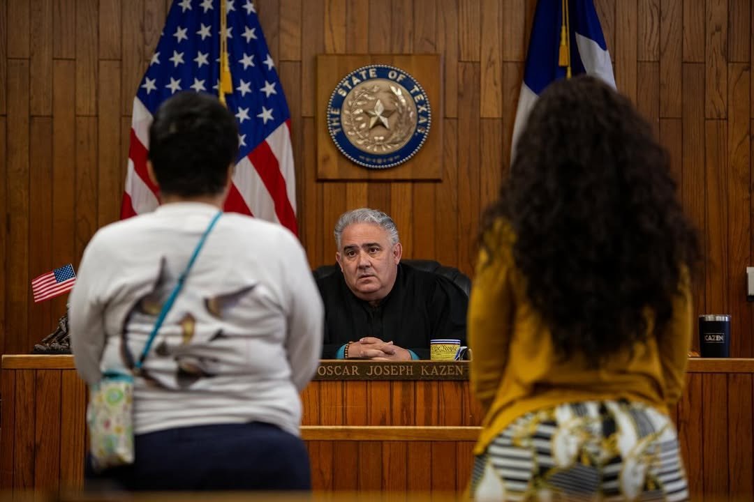 Two women facing a judge in a courtroom with wooden walls, American flags, and a Texas state seal behind the judge's bench.
