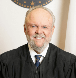 Portrait of an elderly judge in black robe, smiling in a courtroom setting.