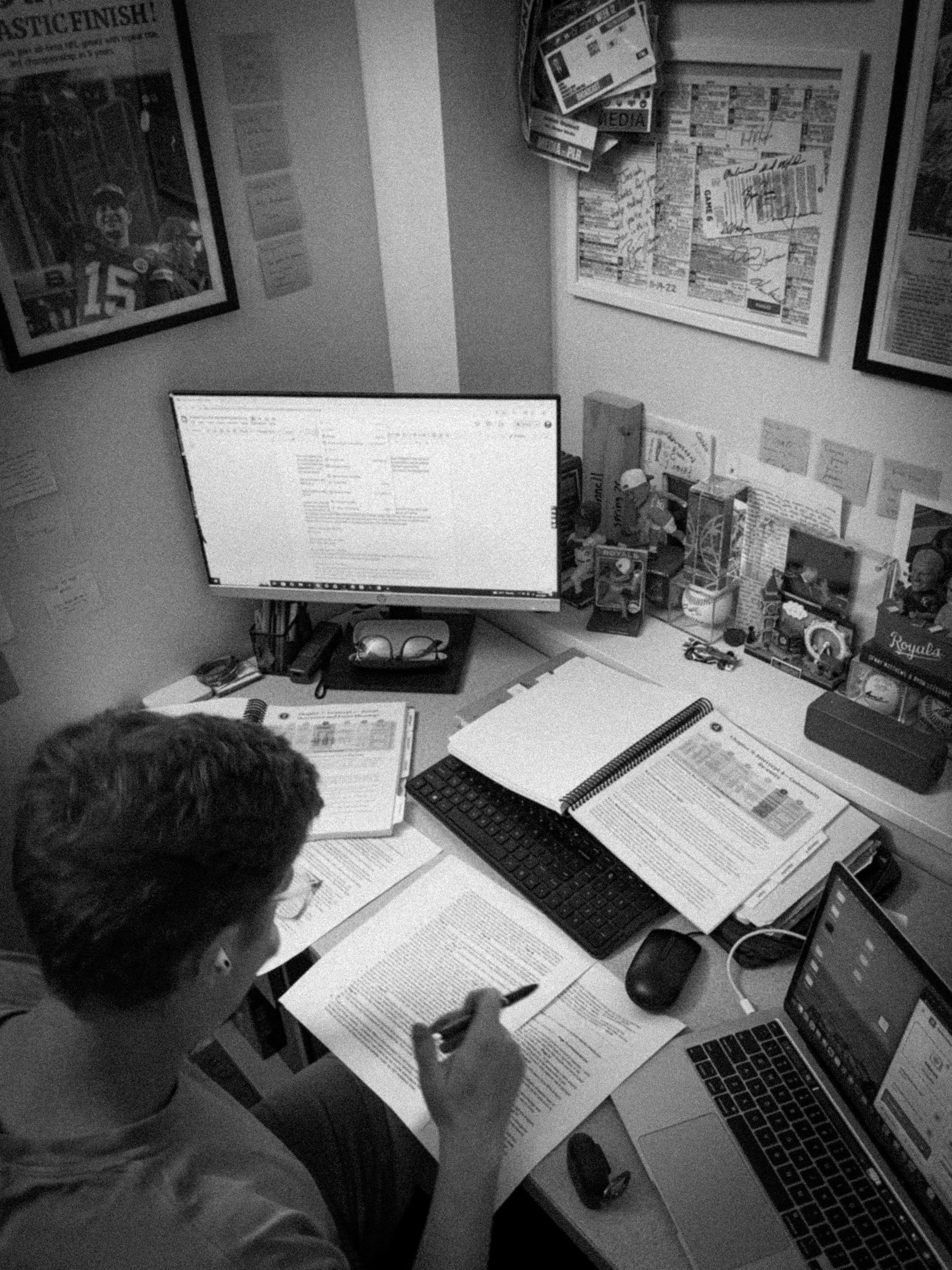 A person is working at a cluttered desk with a computer monitor, papers, books, and office supplies, surrounded by bulletin boards and posters on the walls.
