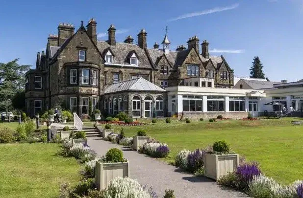 A large historic mansion with a stone exterior and multiple chimneys, surrounded by a well-manicured garden with flower beds and a paved walkway.