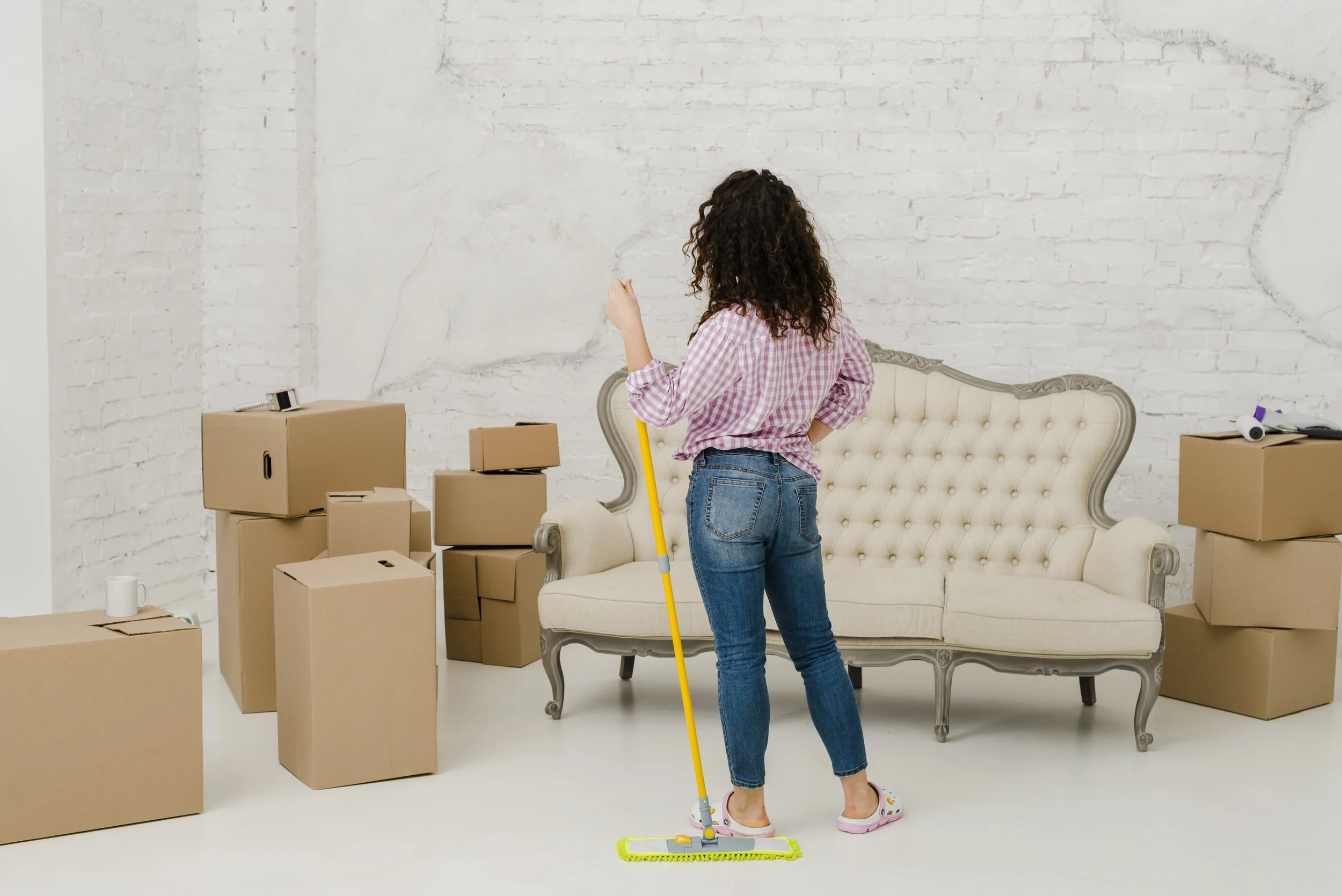 A woman with curly hair in a pink checkered shirt and jeans mops the floor in a room with cardboard boxes and a vintage cream-colored sofa against a white brick wall.