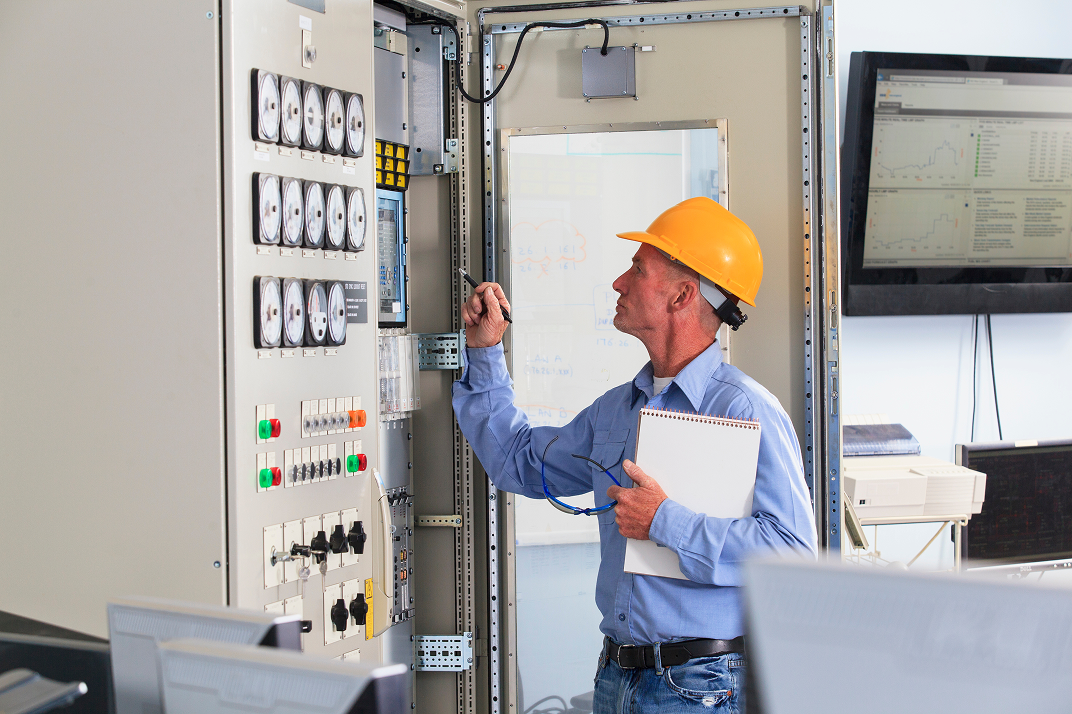 A man wearing a yellow safety helmet and blue shirt inspecting electrical panel with controls and gauges, holding a clipboard and glasses, in a control room with monitors and whiteboard.