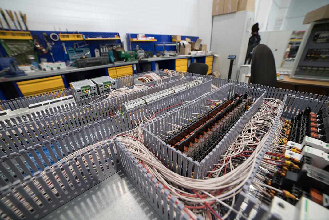 Open electrical control panel with wires, relays, and circuit components inside, located in a workshop or manufacturing space.