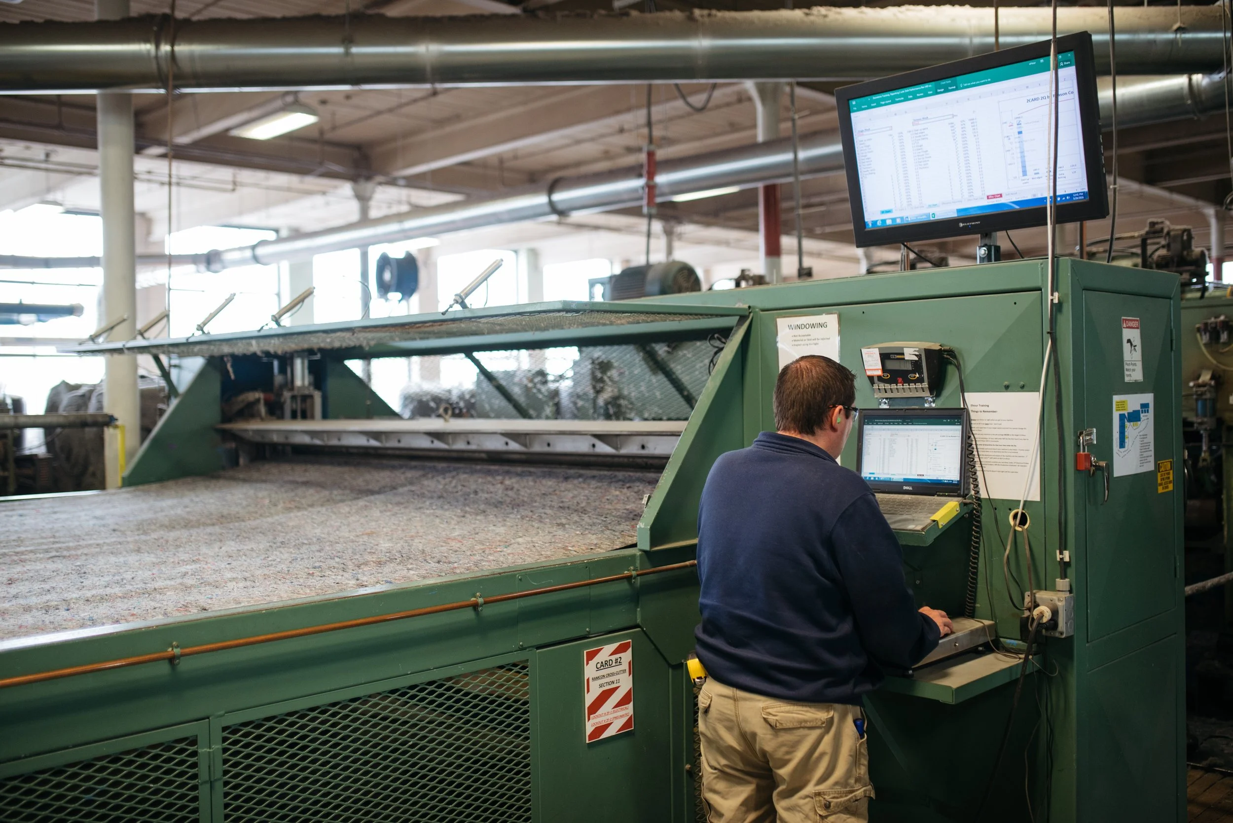 A man working on a computer in an industrial setting next to a large green machine with a monitor overhead displaying data, in a spacious factory or manufacturing plant.