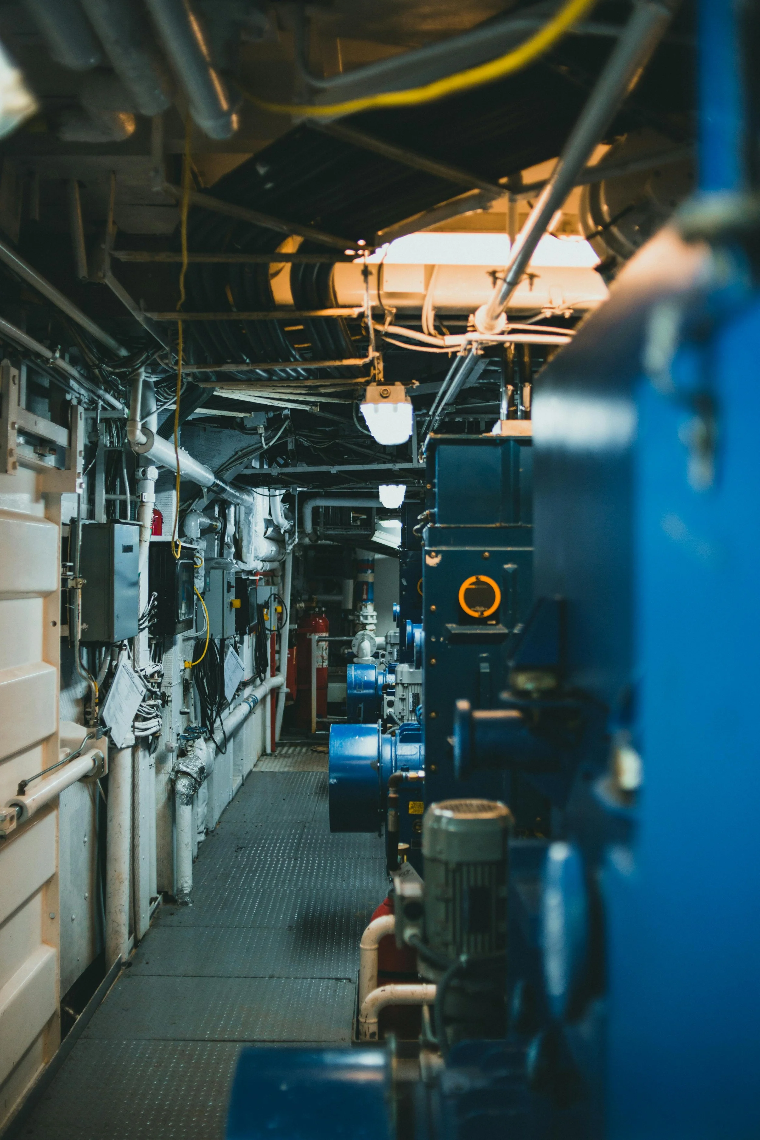 The image shows a corridor of an industrial machine room filled with pipes, valves, and electrical equipment, with a focus on blue machinery on the right and a pathway leading into the background.