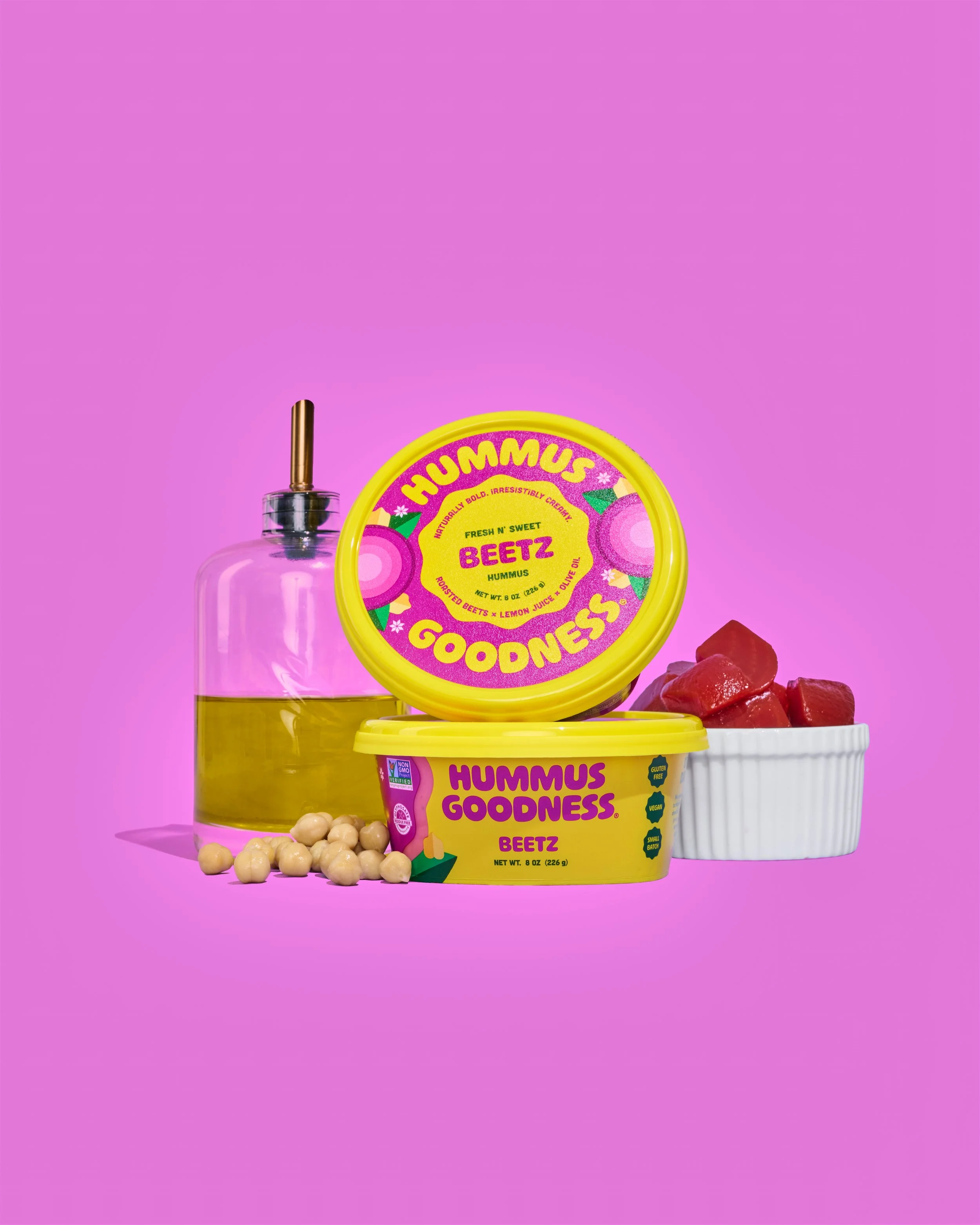 Hummus Goodness Beetz Hummus -Containers of hummus, chickpeas, a glass bottle of olive oil, and a small bowl of diced beets on a purple background.