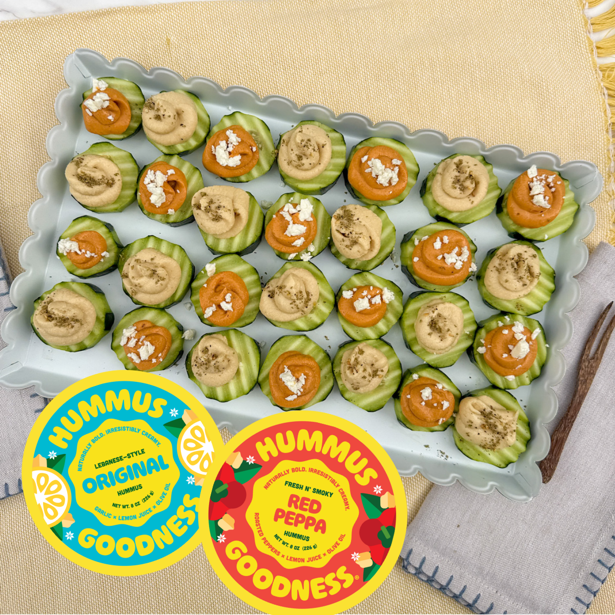 A tray of cucumber slices topped with hummus, red pepper hummus, and sprinkled with herbs, arranged in a neat pattern on a decorative plate. Two hummus labels, one for original hummus and one for red pepper hummus, are placed in front of the tray.