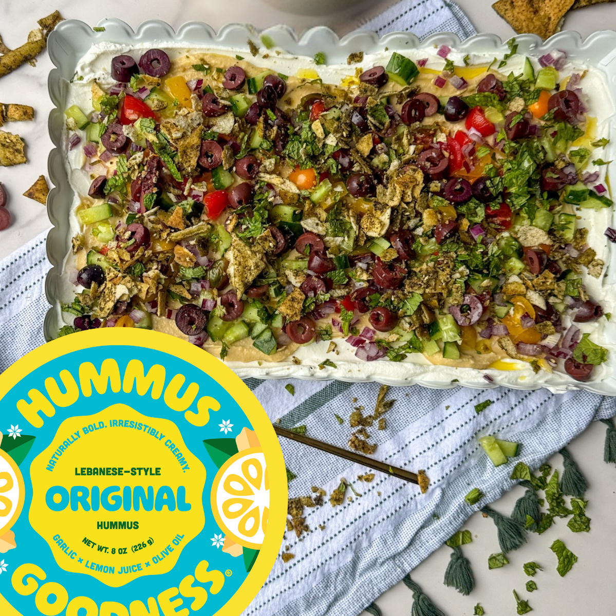 A large rectangular dish of hummus topped with chopped green celery, red tomatoes, purple olives, chopped herbs, and crushed garlic crackers. A hummus label is visible in the bottom left corner.