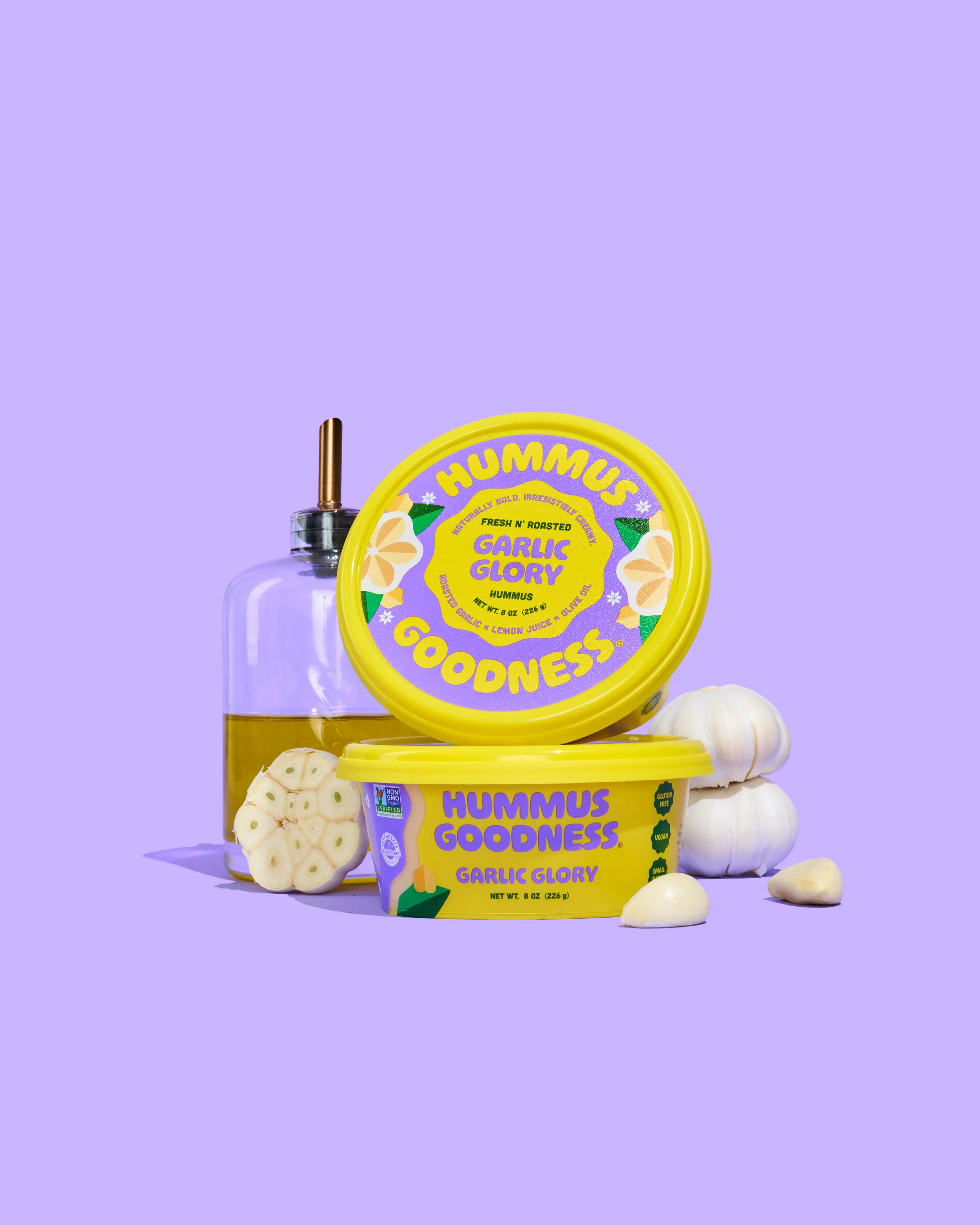 Hummus Goodness Garlic Glory Hummus - Hummus containers labeled 'Garlic Glory' with garlic cloves, a garlic bulb, and a bottle of olive oil on a purple background.