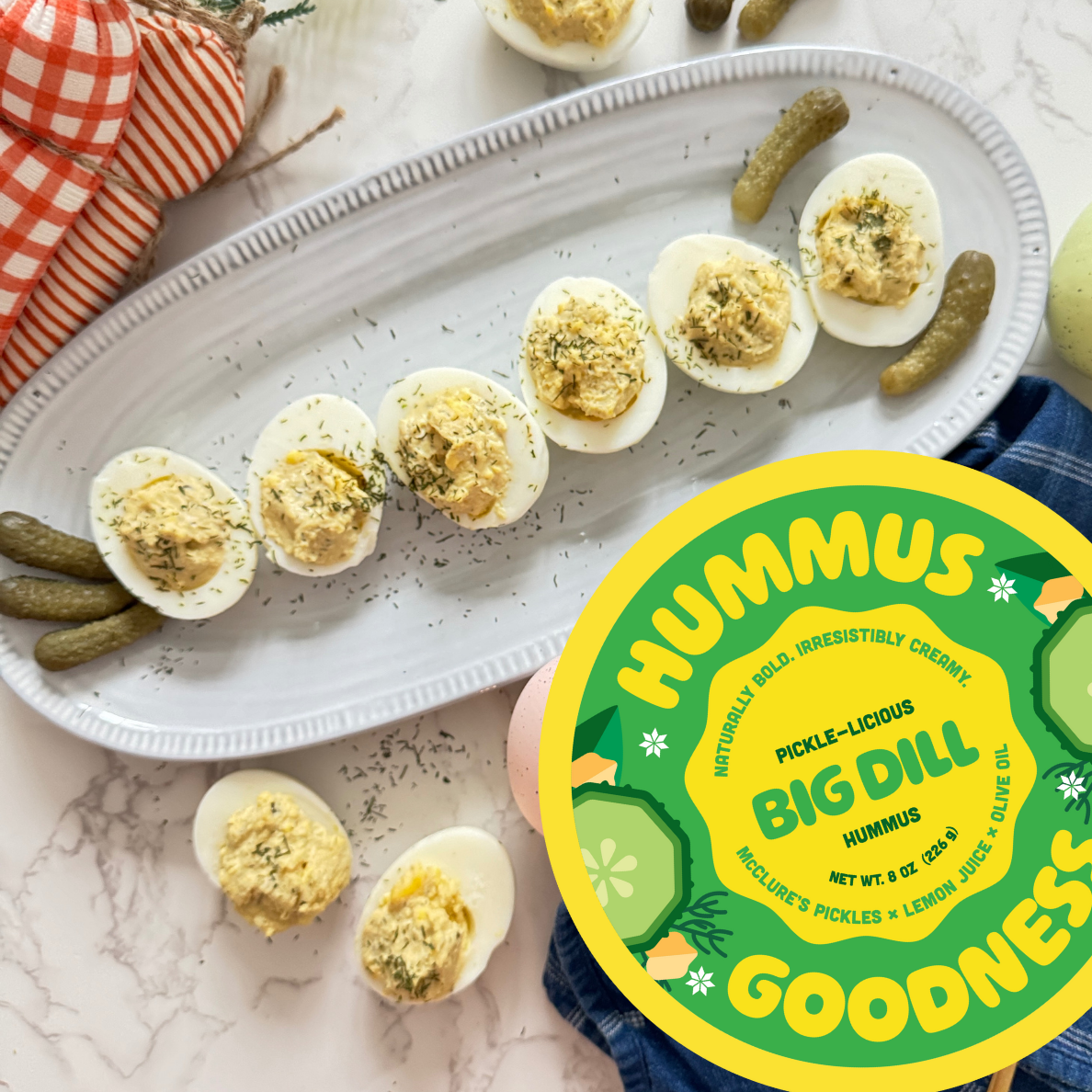 Half-sliced hard-boiled eggs topped with hummus on a white platter, garnished with herbs and small pickles around the eggs, with a container of hummus labeled 'Big Dill' in the foreground.