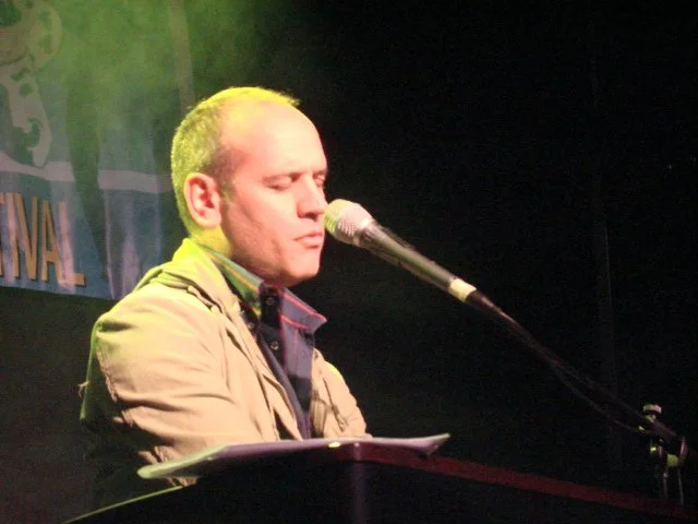 Un uomo che canta o parla al microfono, con una postura seria, durante un'esibizione o un discorso, in un ambiente con sfondo scuro e luci verdi.