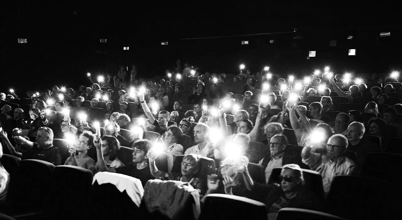 Pubblico seduto in sala con luci del cellulare accese, probabilmente durante un evento o un cinema, in bianco e nero.