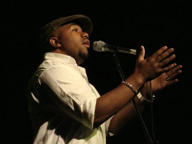 Un uomo che canta o parla in un microfono su uno sfondo nero.