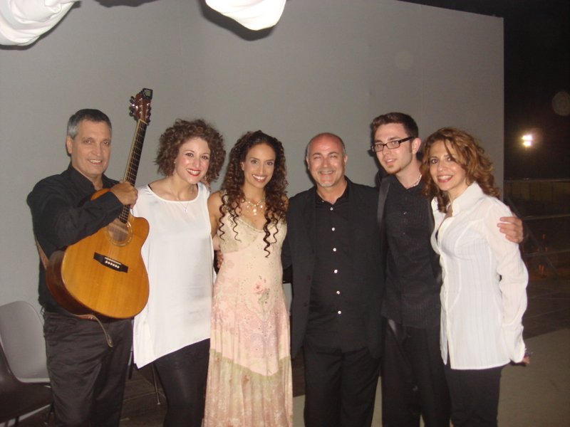 Gruppo di sei persone sorridenti che posano insieme, con uno di loro che tiene una chitarra, in un ambiente interno con parete grigia