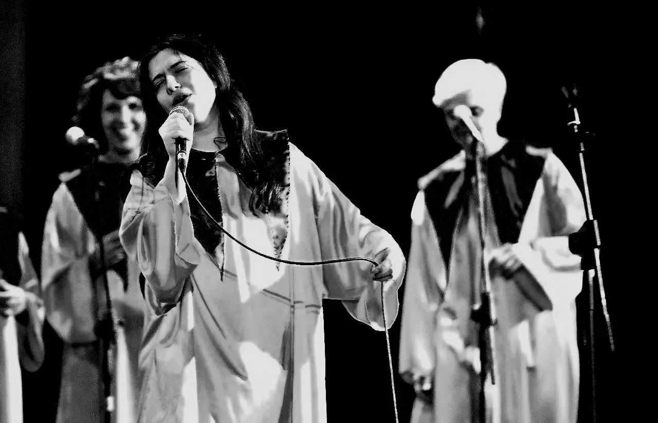 Tre persone cantano con microfoni vestiti con abiti a maniche larghe e colorati, scena in bianco e nero.