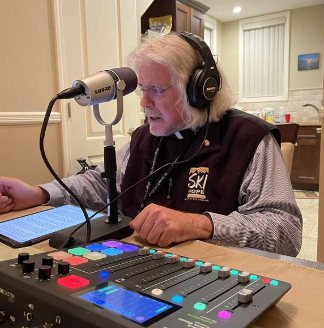 An older man with long white hair and glasses recording in a home studio, wearing headphones and speaking into a microphone, with a digital music controller in front of him.