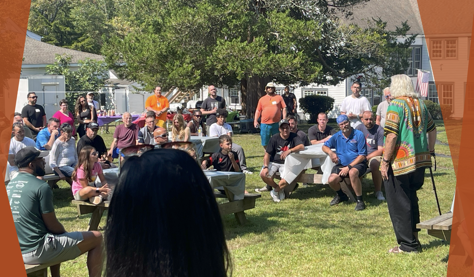 Group of people gathered outdoors in a park for an event, with some seated at picnic tables and others standing, listening to a speaker with white hair, wearing a colorful shirt, holding a bag, under a large tree.