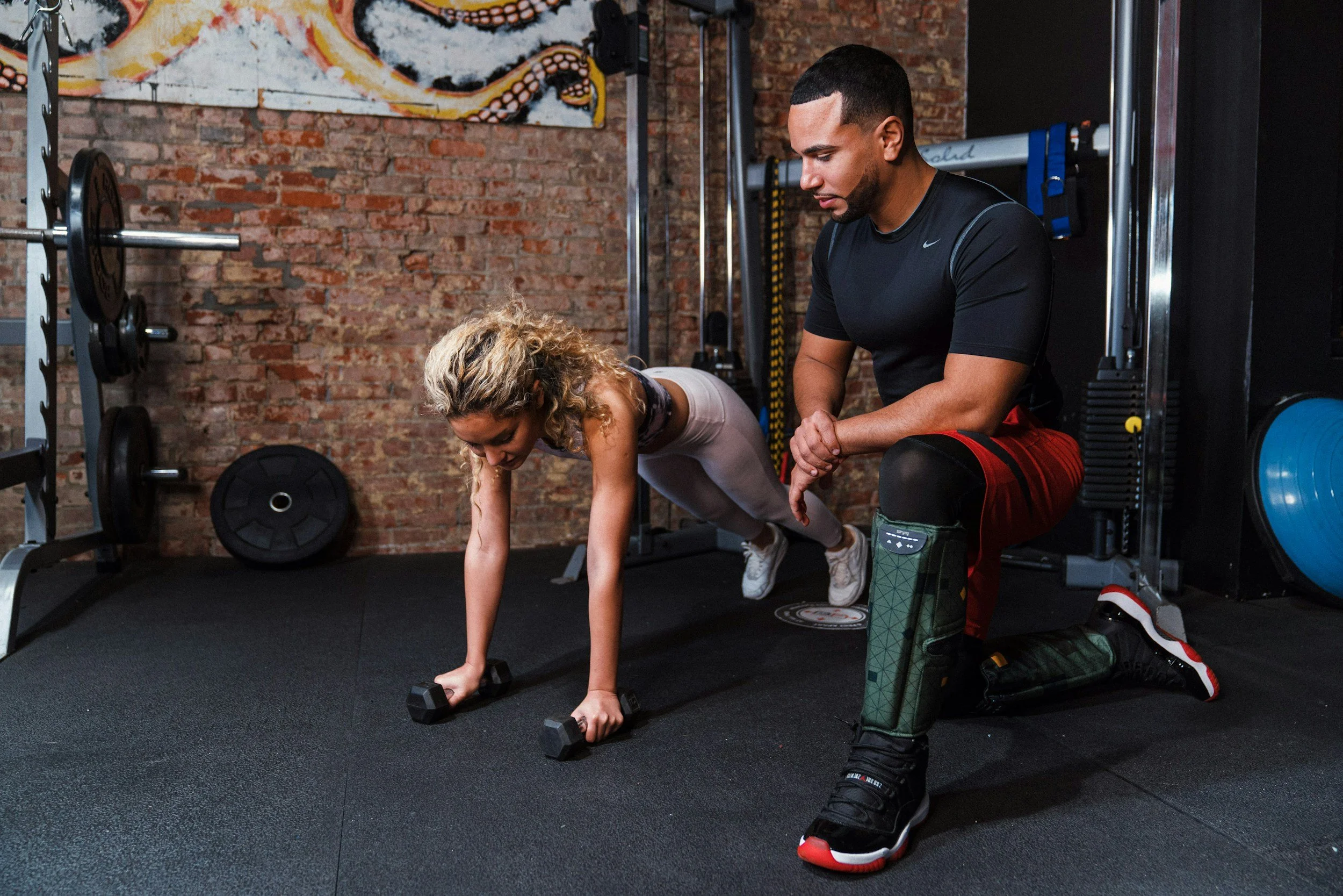 A man in athletic clothing kneeling on one knee next to a woman with curly blonde hair who is doing push-ups with dumbbells in a gym. The man is observing her and appears to be coaching or encouraging her. The gym has brick walls, weight lifting equipment, and a spider painting on the wall.