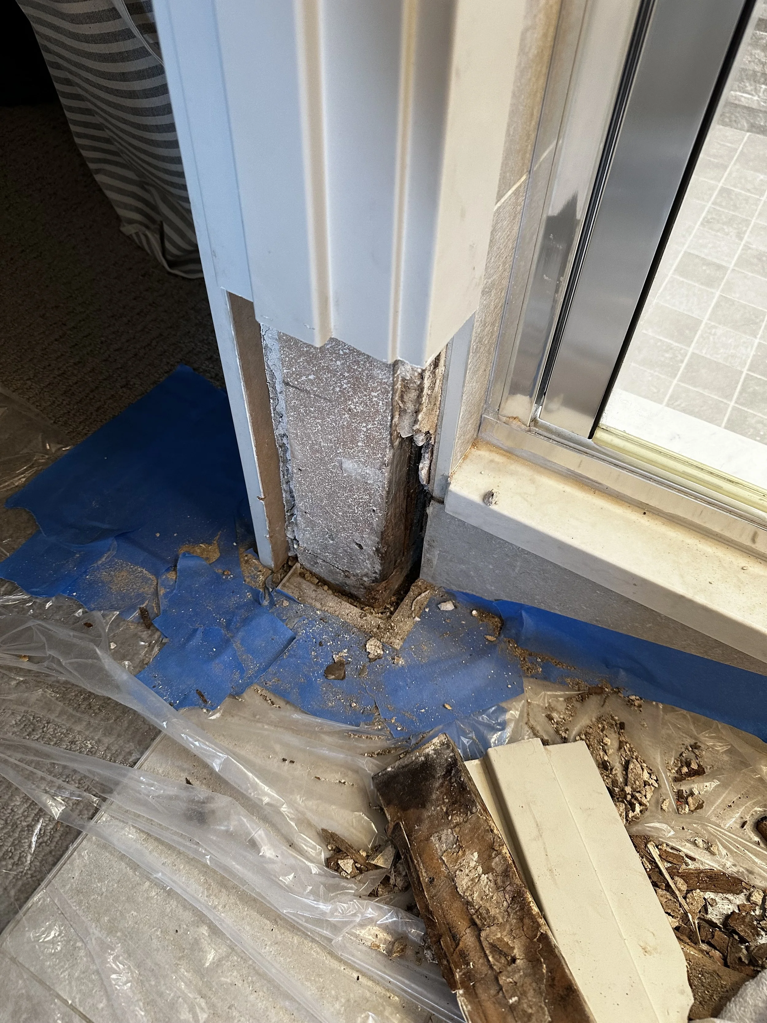 Close-up of a building foundation repair site near a sliding glass door, with exposed concrete and rusted metal reinforcement, surrounded by blue painter's tape and debris on the floor.