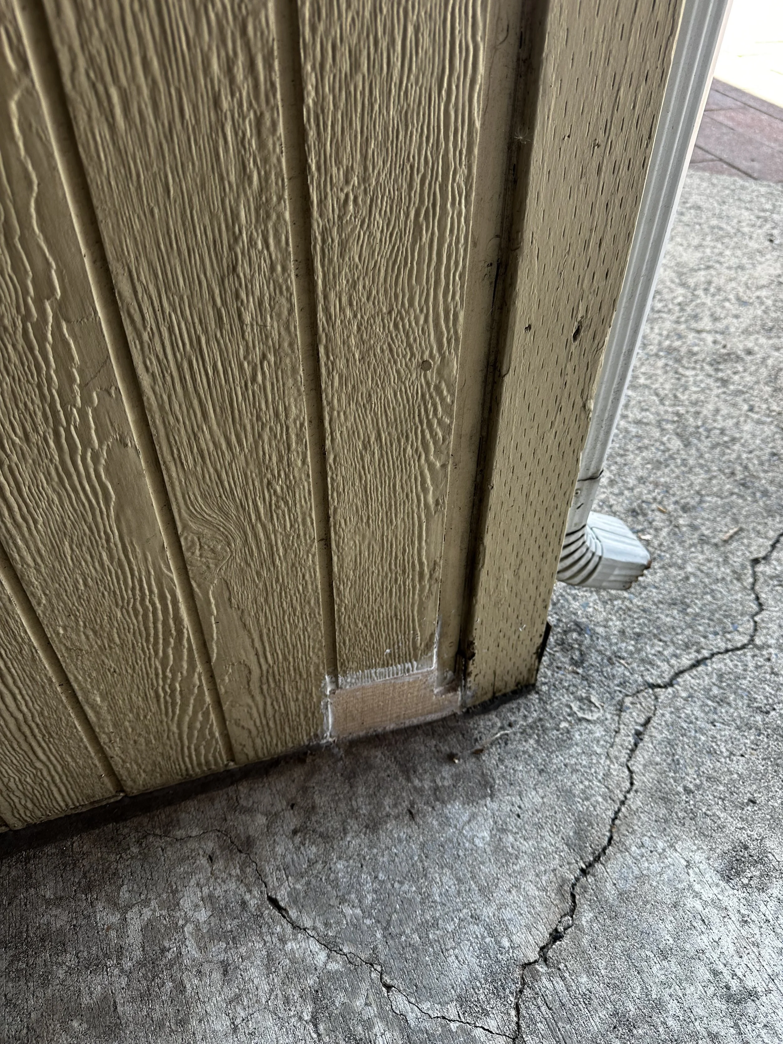Close-up of a corner of a yellow wooden building with textured vertical siding, showing a small area of white paint or caulk and a crack in the concrete ground.