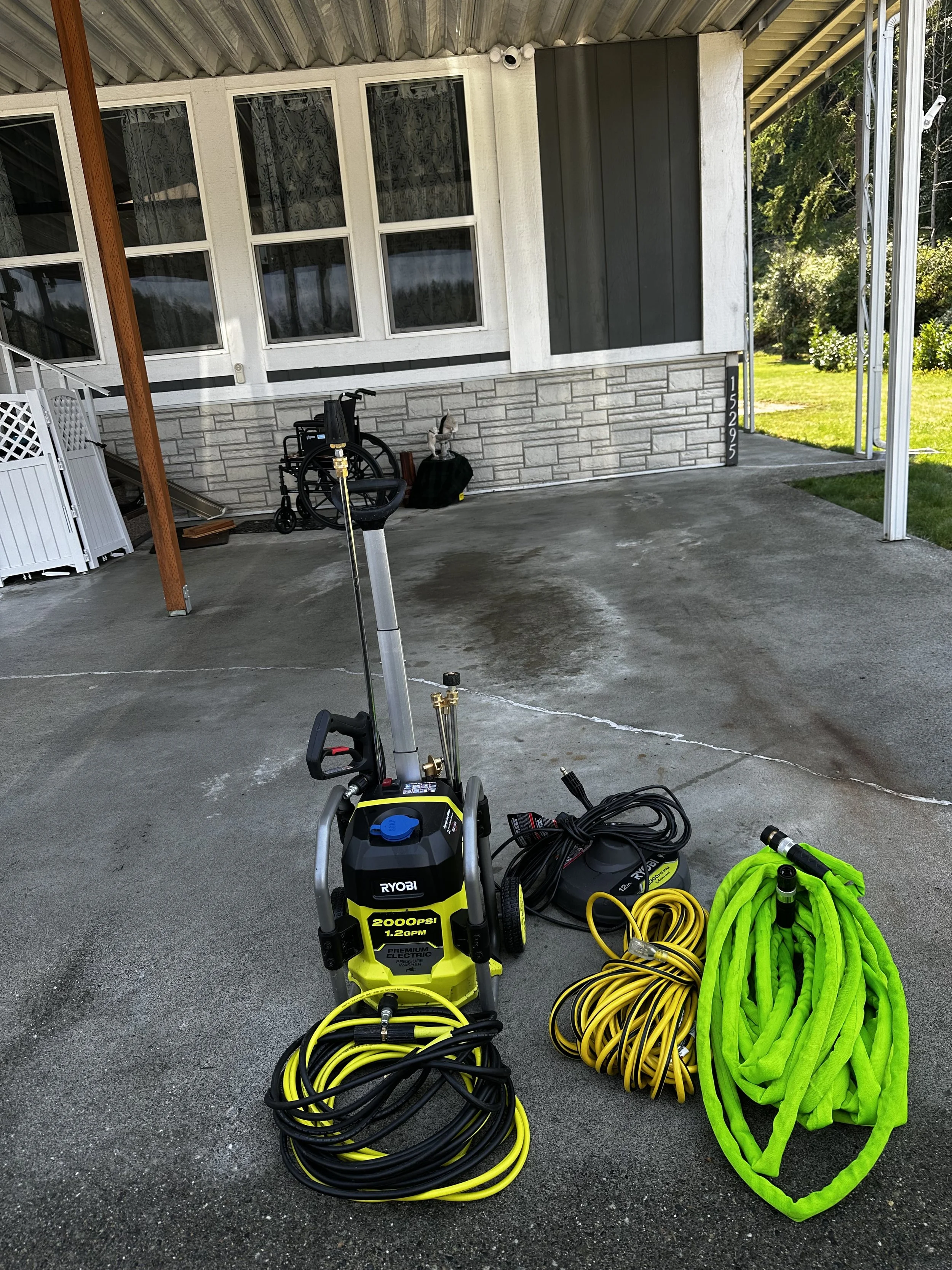 Pressure washer with black and yellow design, hoses, and extension cord on a concrete driveway in front of a house with grey and white siding, a stone foundation, and windows.