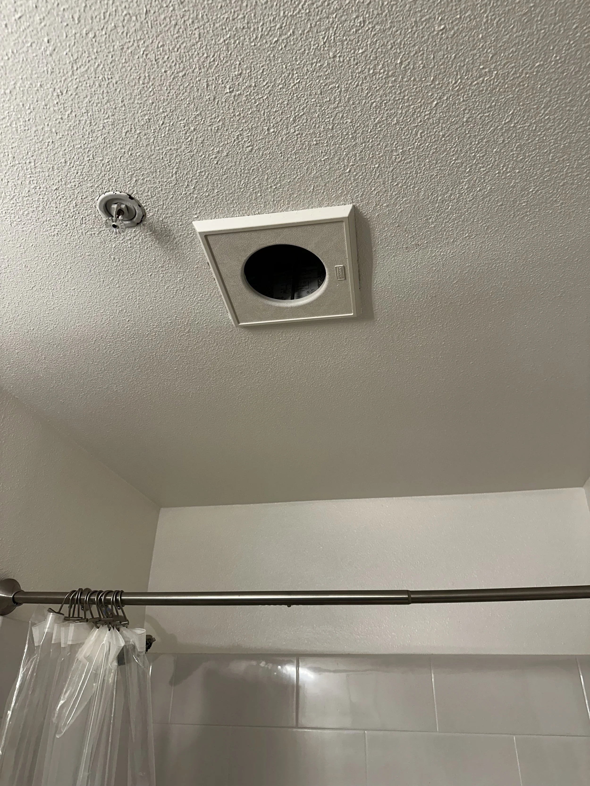 Ceiling vent above a shower curtain rod, which has a plastic shower curtain, in a bathroom.