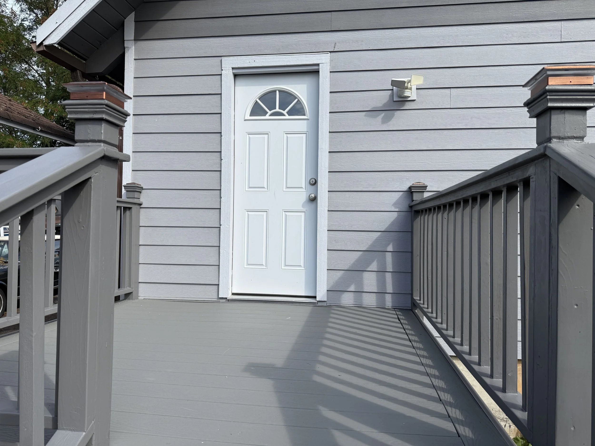Front porch with gray wooden railing and gray siding, white door with small semi-circular window, outdoor light fixture, and steps leading up to the door.