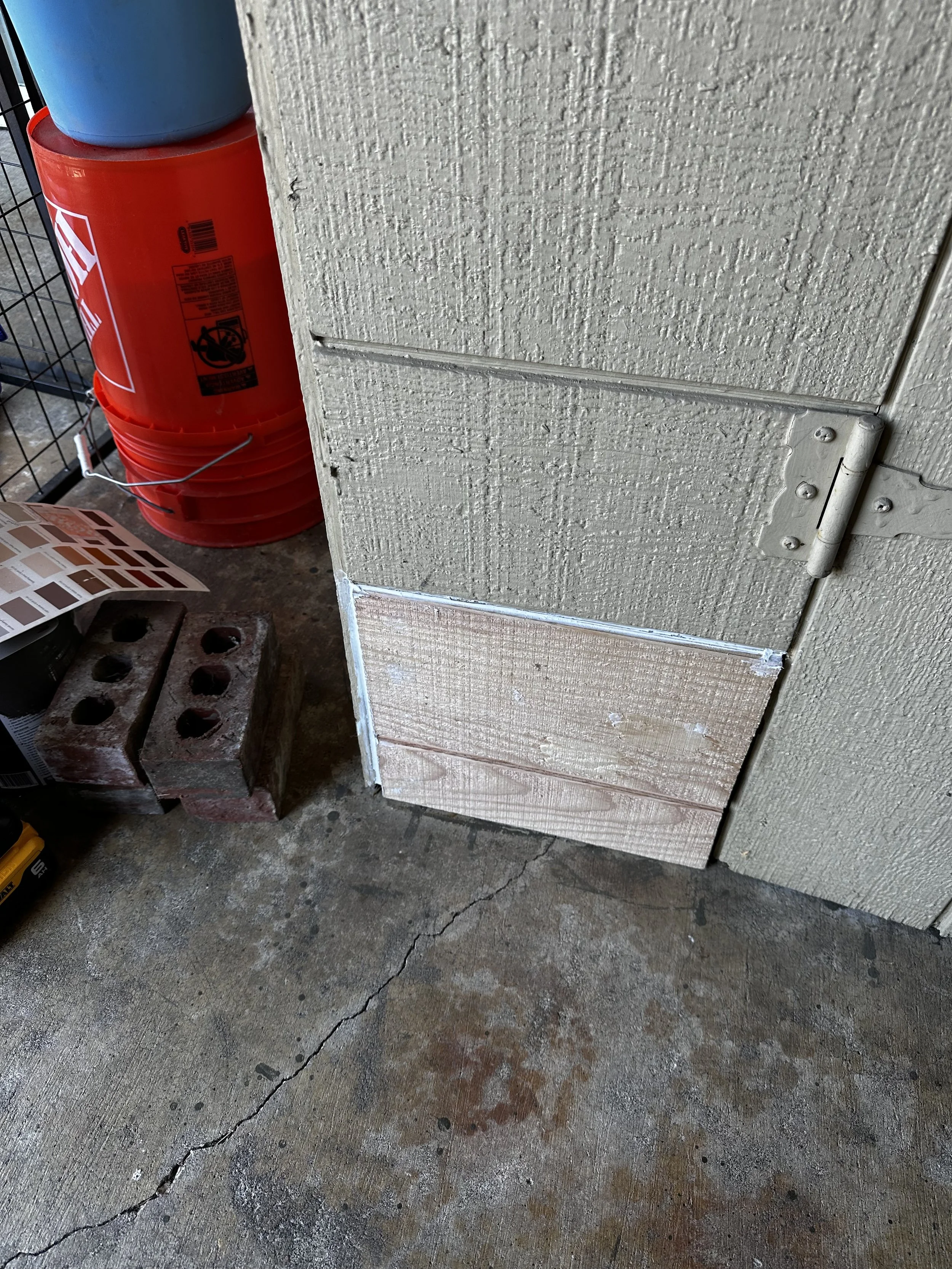 Close-up of a garage floor with a crack, showing a wooden board leaning against a textured beige wall, with a red safety barrel, a blue bucket, bricks, and paint sample sheet nearby.