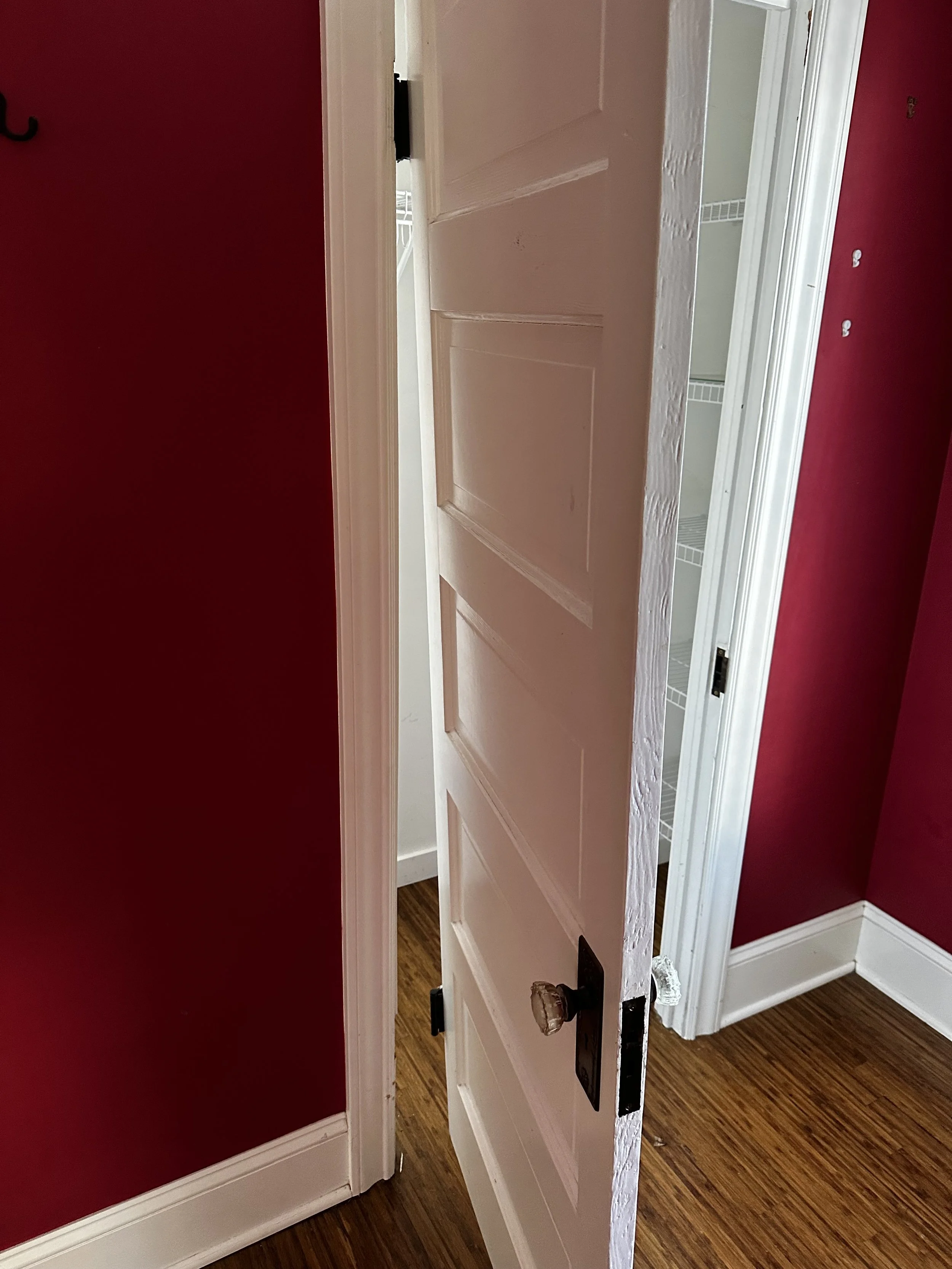 Partially open white door revealing a closet with white wire shelves, hardwood floor, red walls, and white trim.