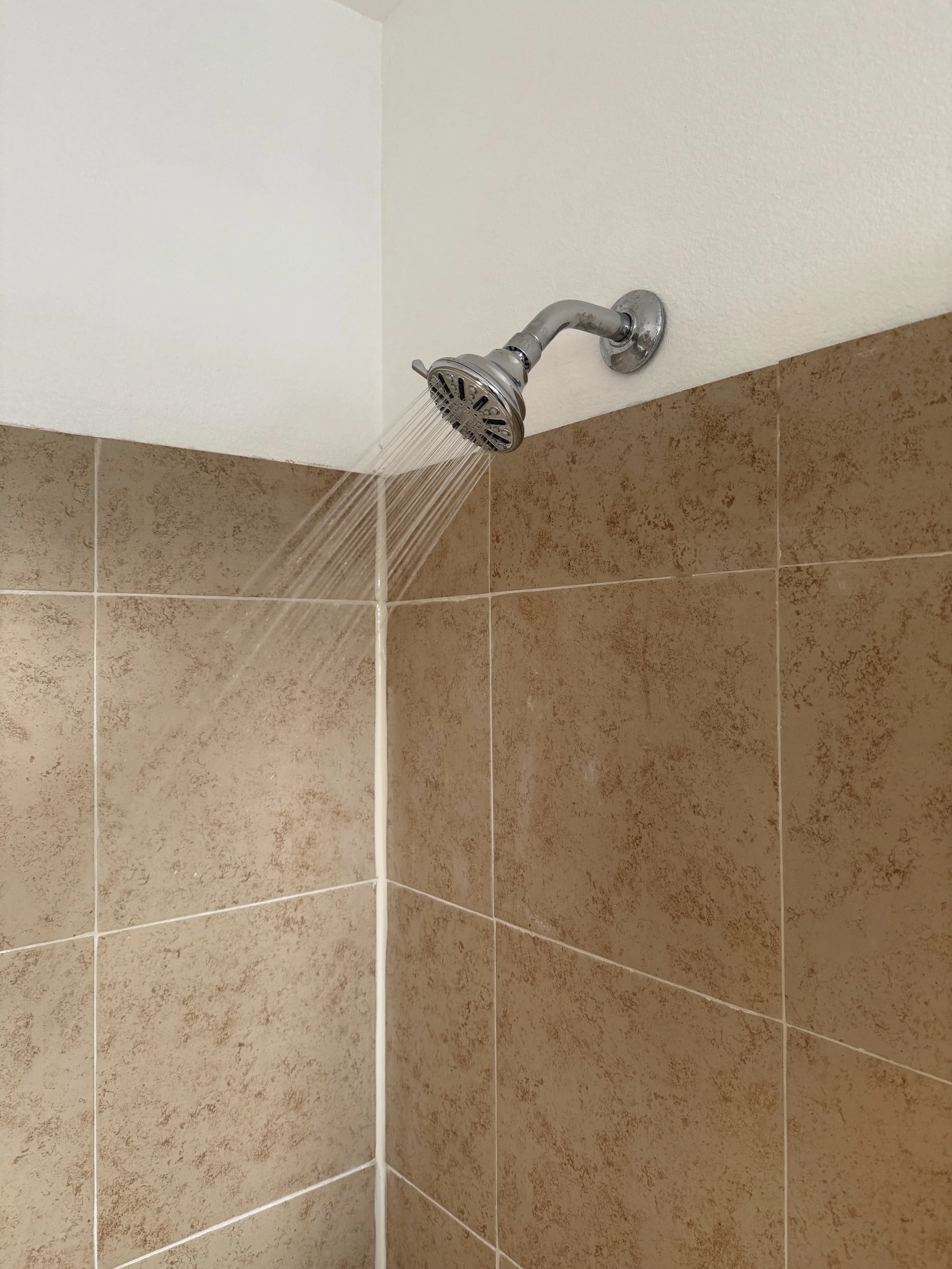 A showerhead mounted on a wall with tan tiles, water flowing from it.
